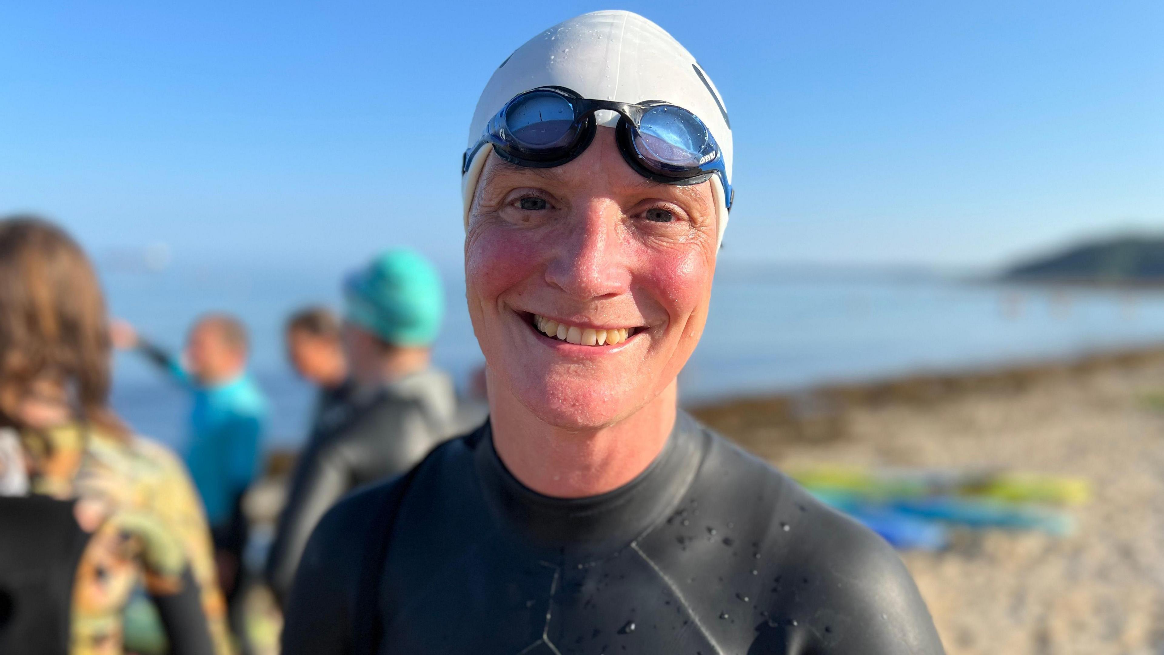 A woman in a wet suit, swim hat and goggles smiles for the camera 