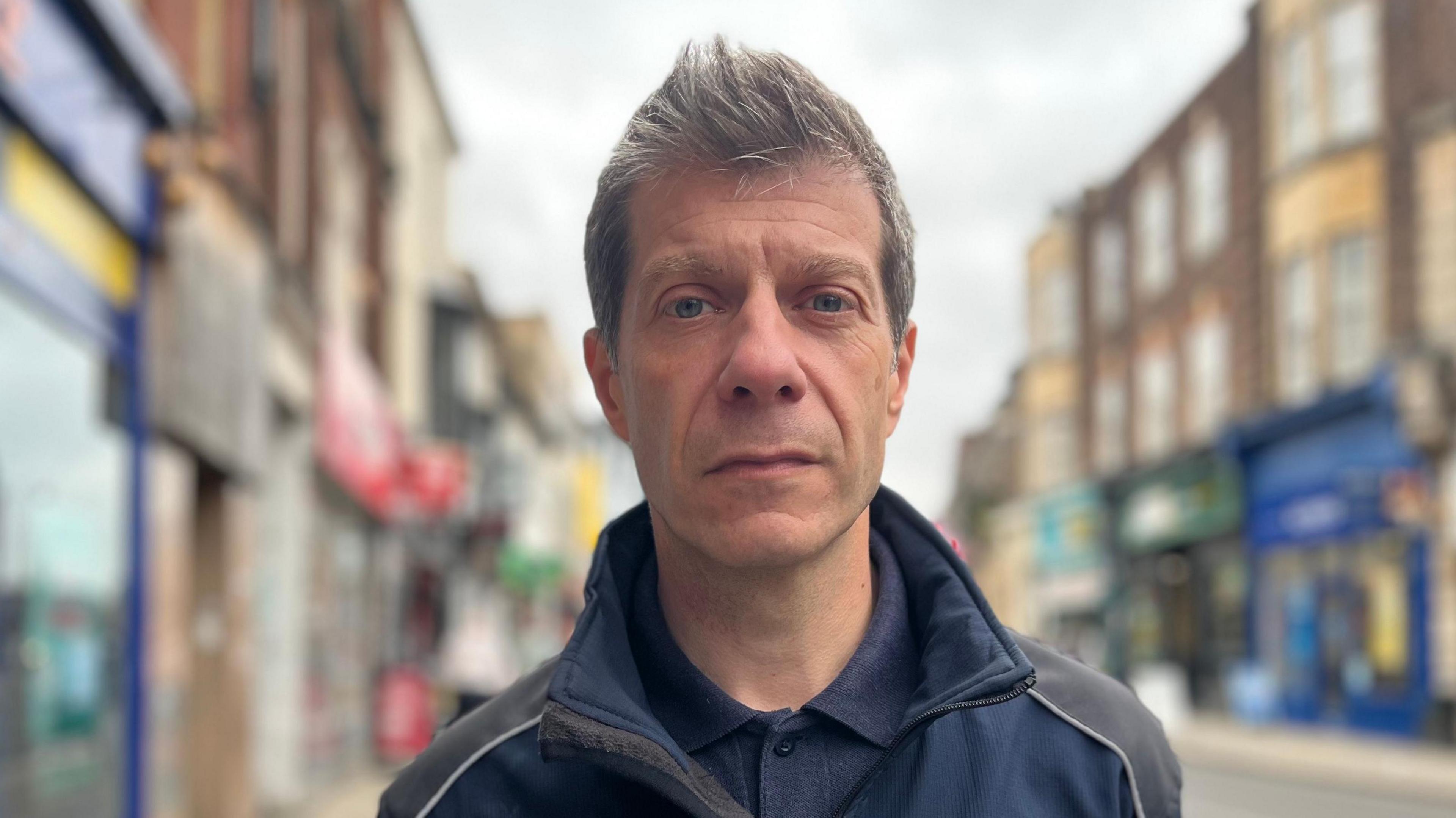 Pep Cipriano is photographed outside on a street with a number of shopfronts behind him. He is looking directly at the camera with a solemn expression. He has grey hair and is wearing a blue shirt and jacket. 