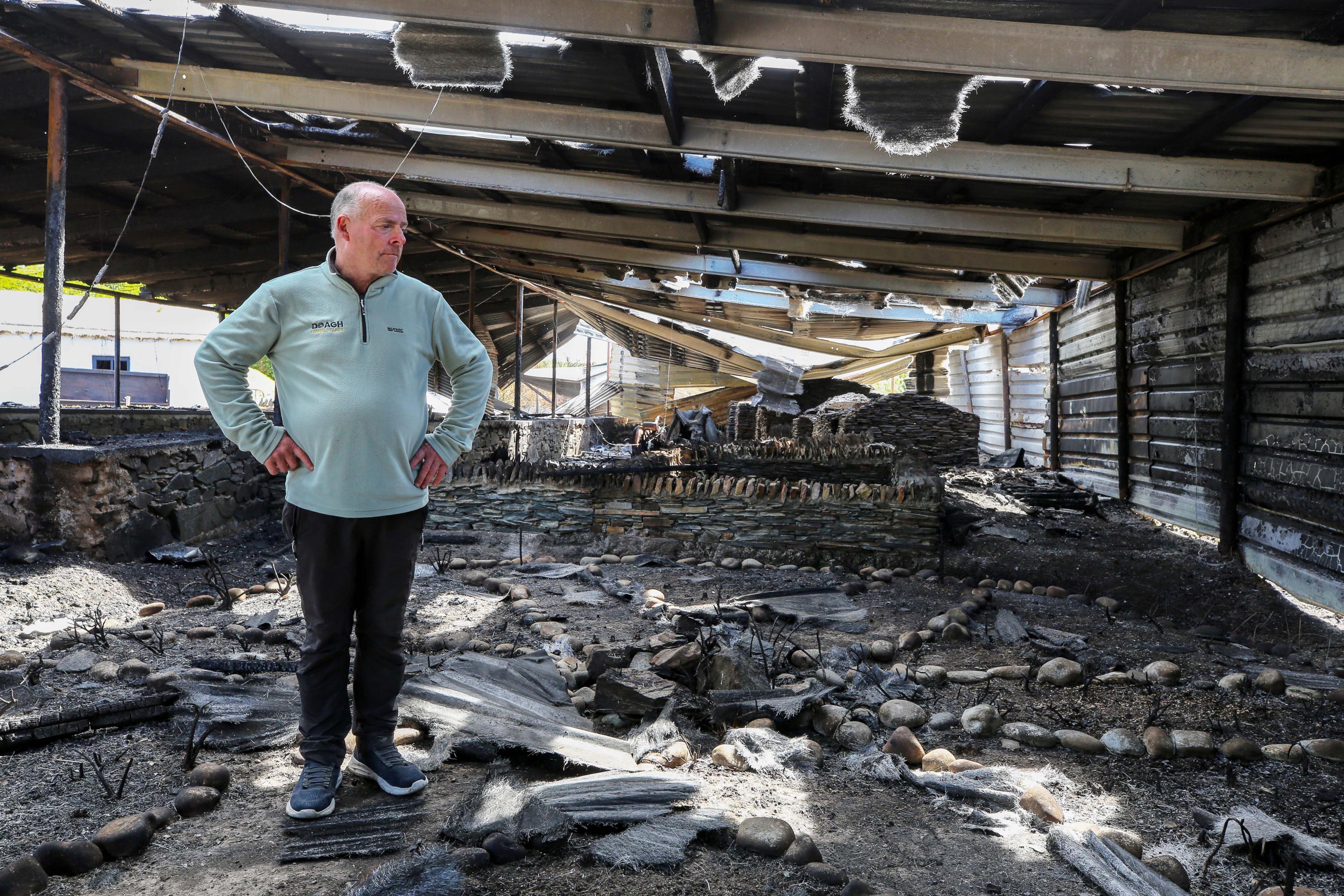 A man wearing a blue jumper stands in the wreckage of a fire. he has his hands on his hips and stares into the devastation. All around his are charred remains of what the museum once was.