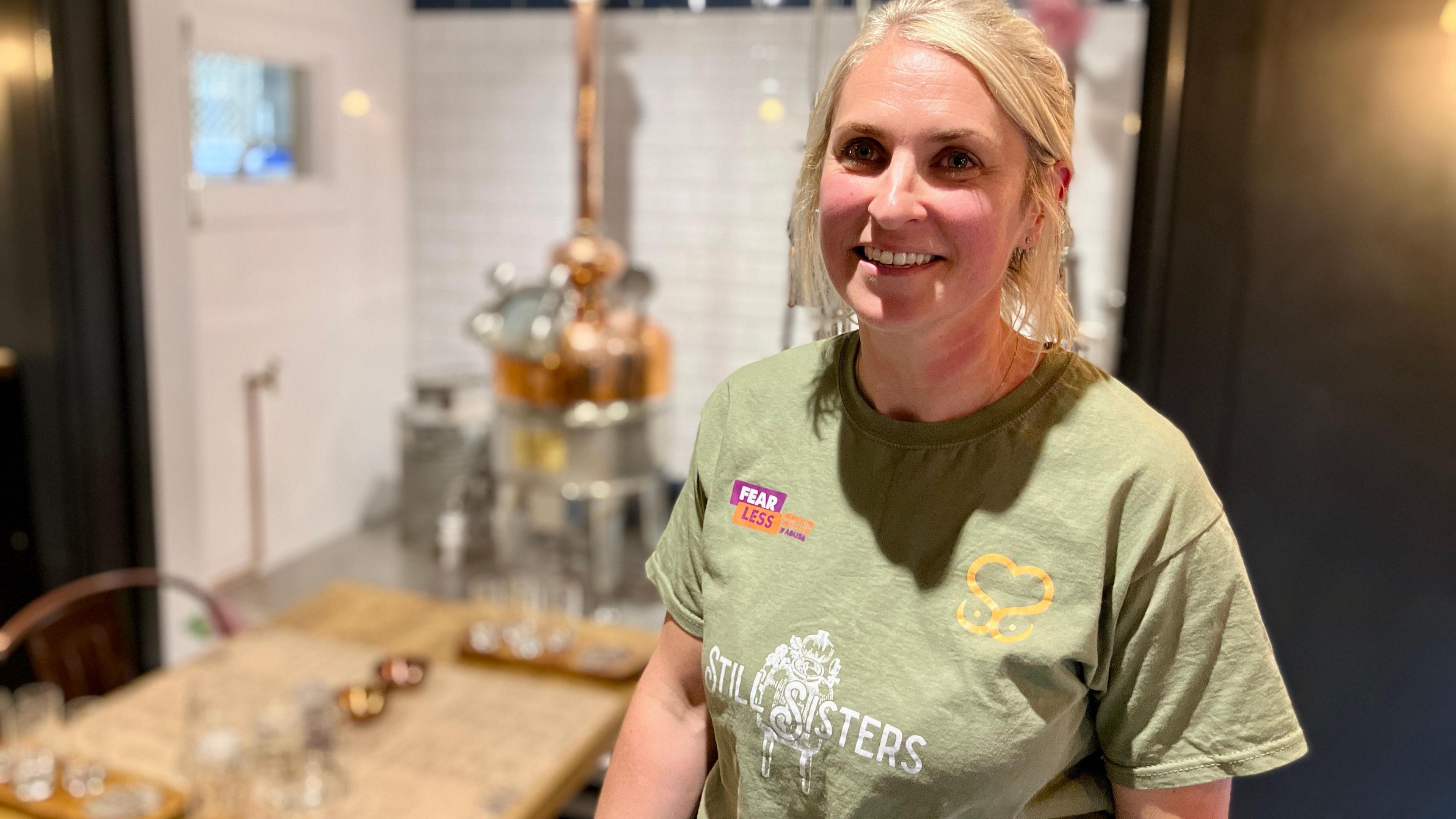 Tori Jordan wearing a green t-shirt with blonde hair smiling at teh camera with her distillery in the background, blurred out