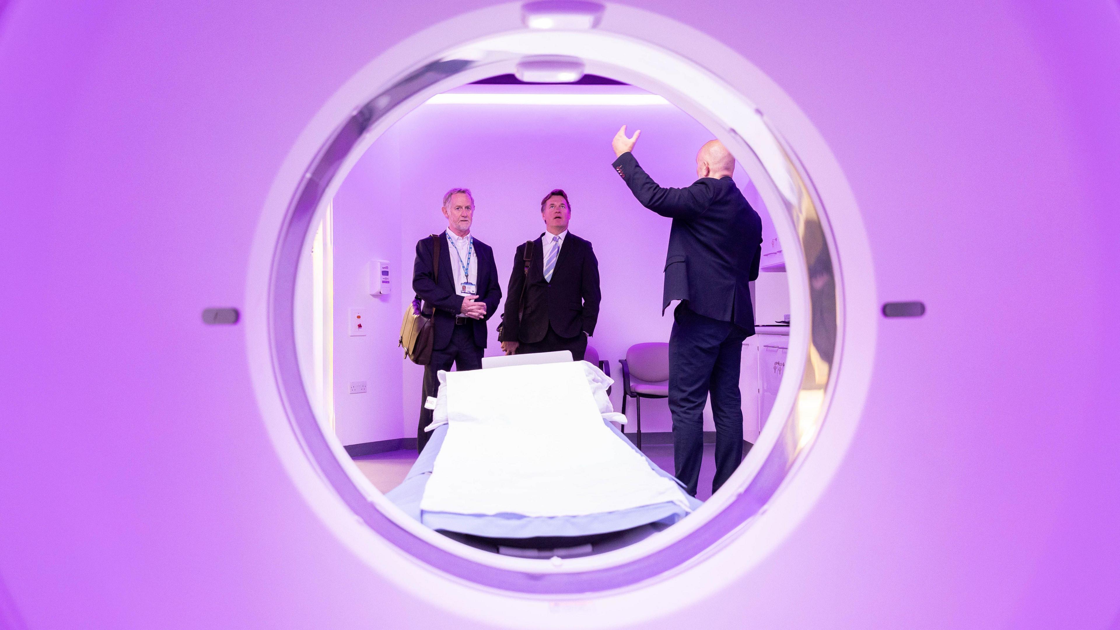 Three men stand in a hospital room by a hospital bed. The image is taken through a purple circular fixture.