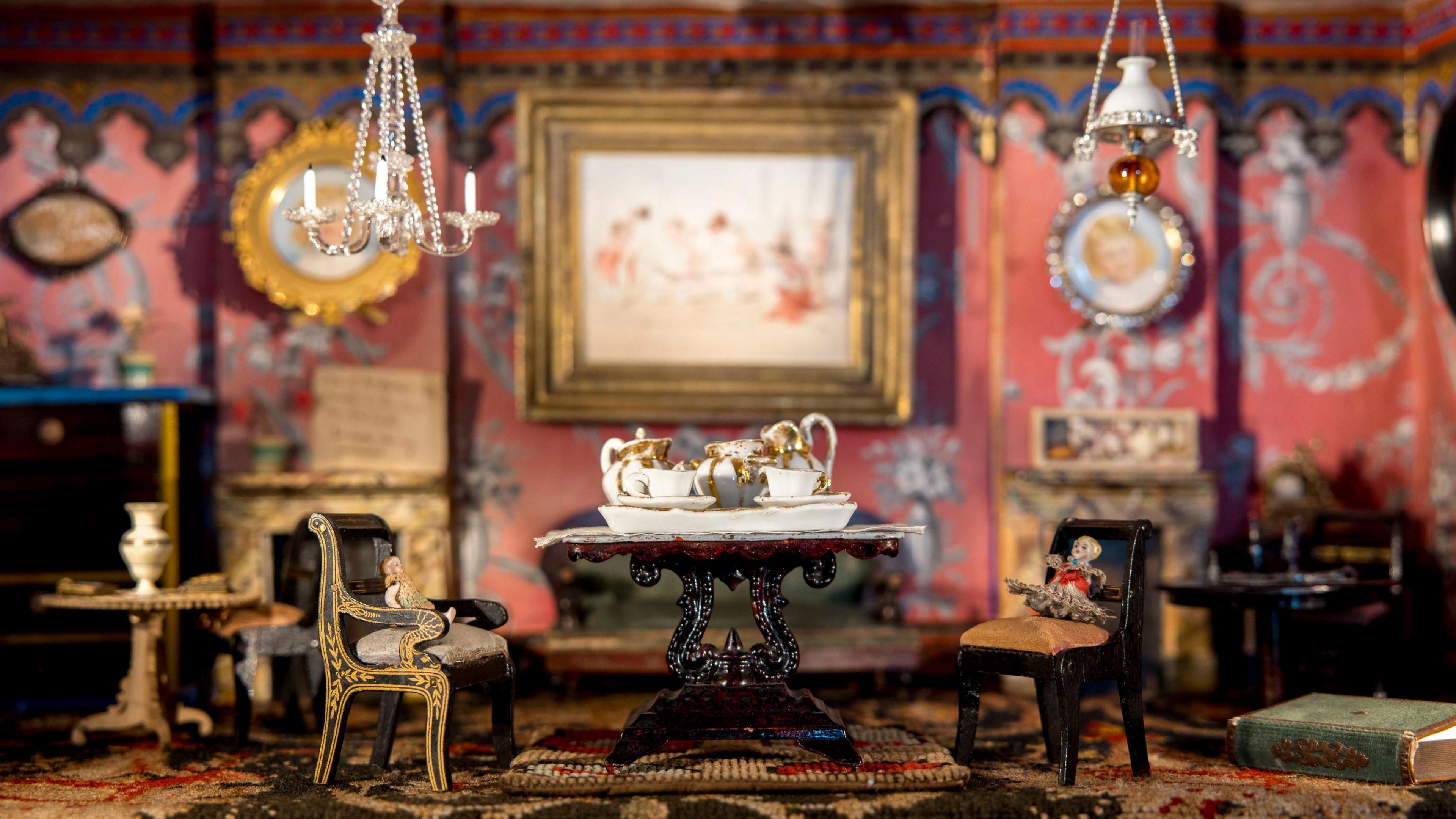 A close-up of one of the rooms. It has pink, patterned wallpaper with a table and two chairs in the centre. Pictures hang on the wall in gold frames and chandeliers hang from the ceiling.