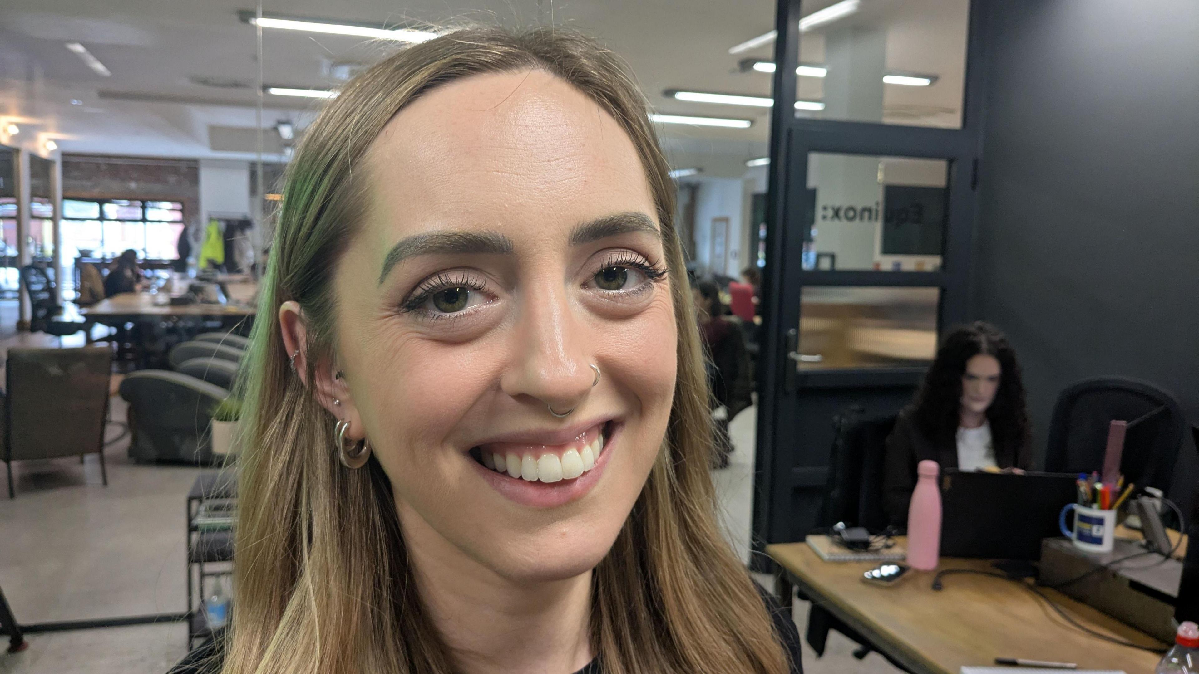 Caroline smiles at the camera. She has medium length blond hair, has two nose piercings and multiple earrings. A large office is visible behind her. It is a head shot of her. 