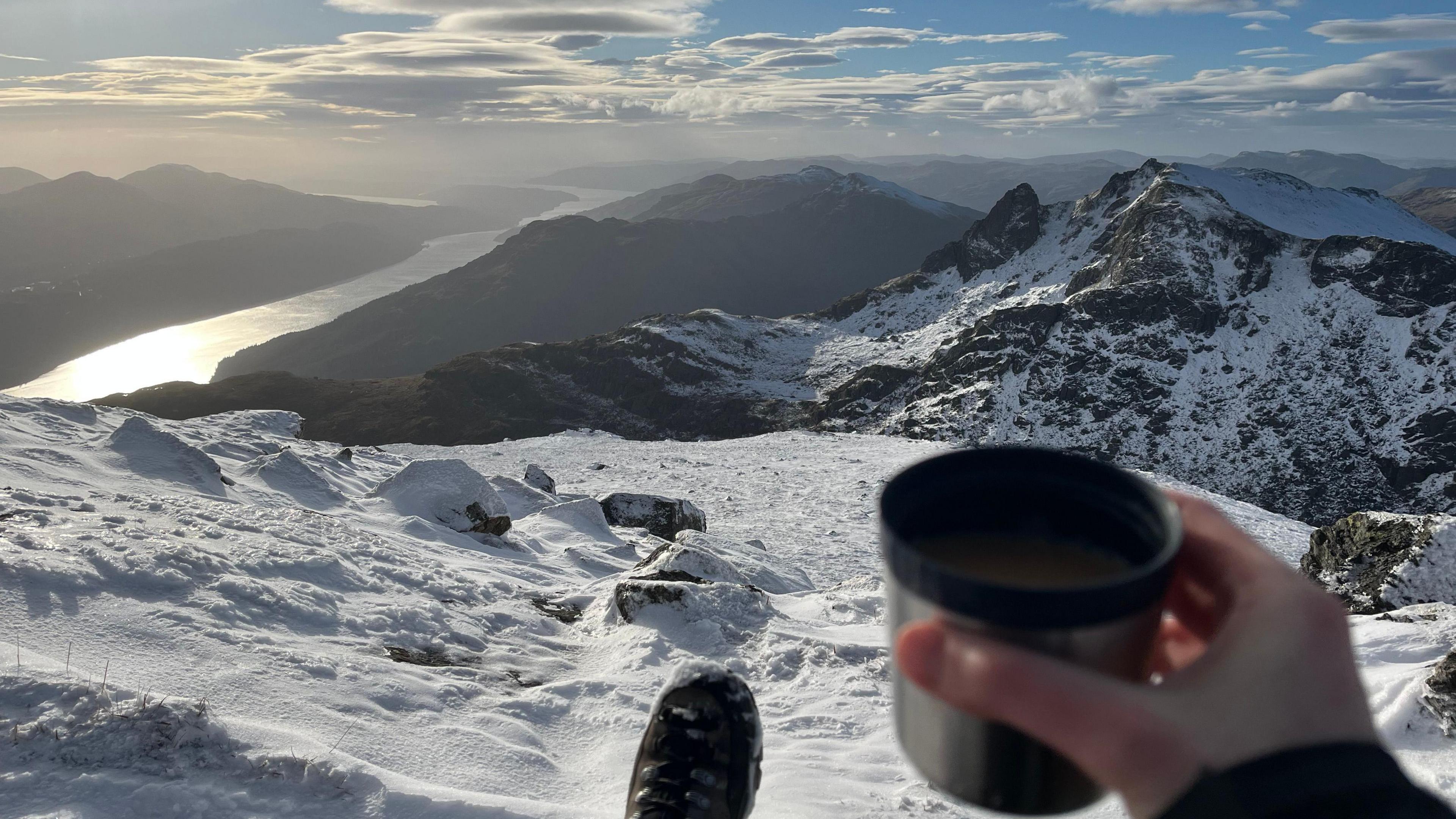 A hand holds a cup overlooking snow-covered peaks and a bright sky.