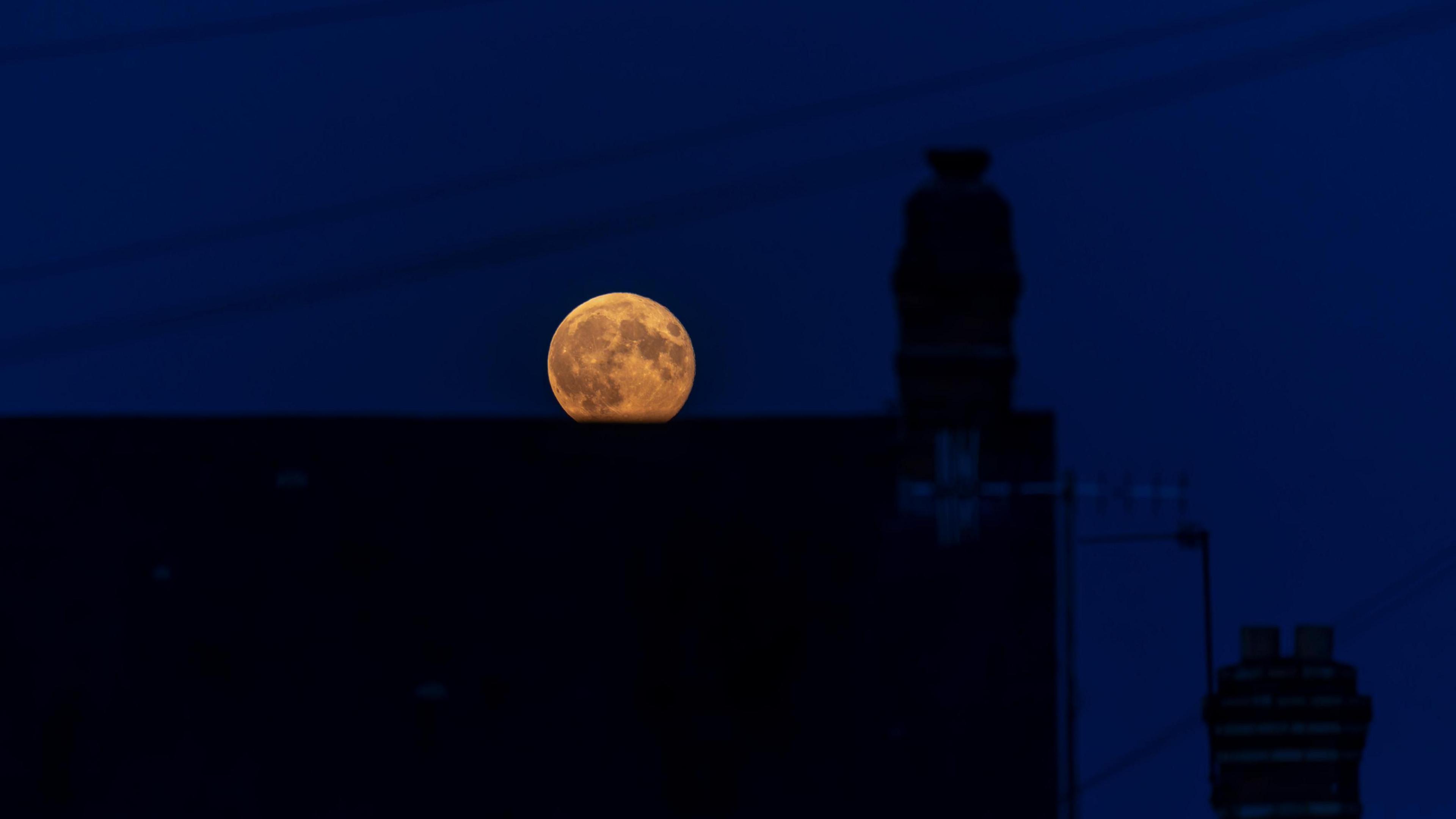 The orange-looking moon is partly obscured by a silhouetted building. The sky is a deep shade of blue. 