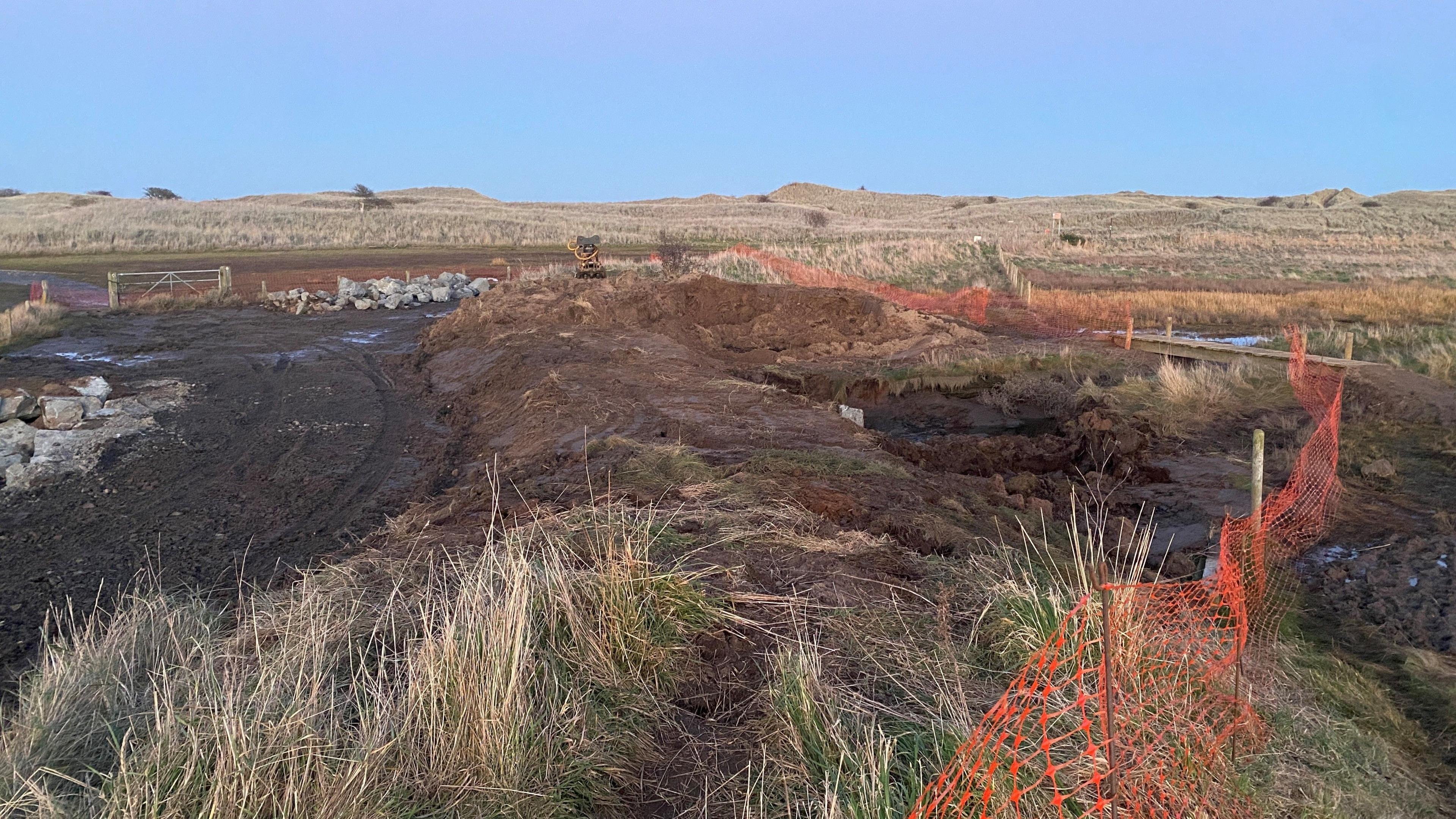 An area of dunes has a muddy track passing through it with a raised bed of earth. To the right is an orange plastic temporary fence.