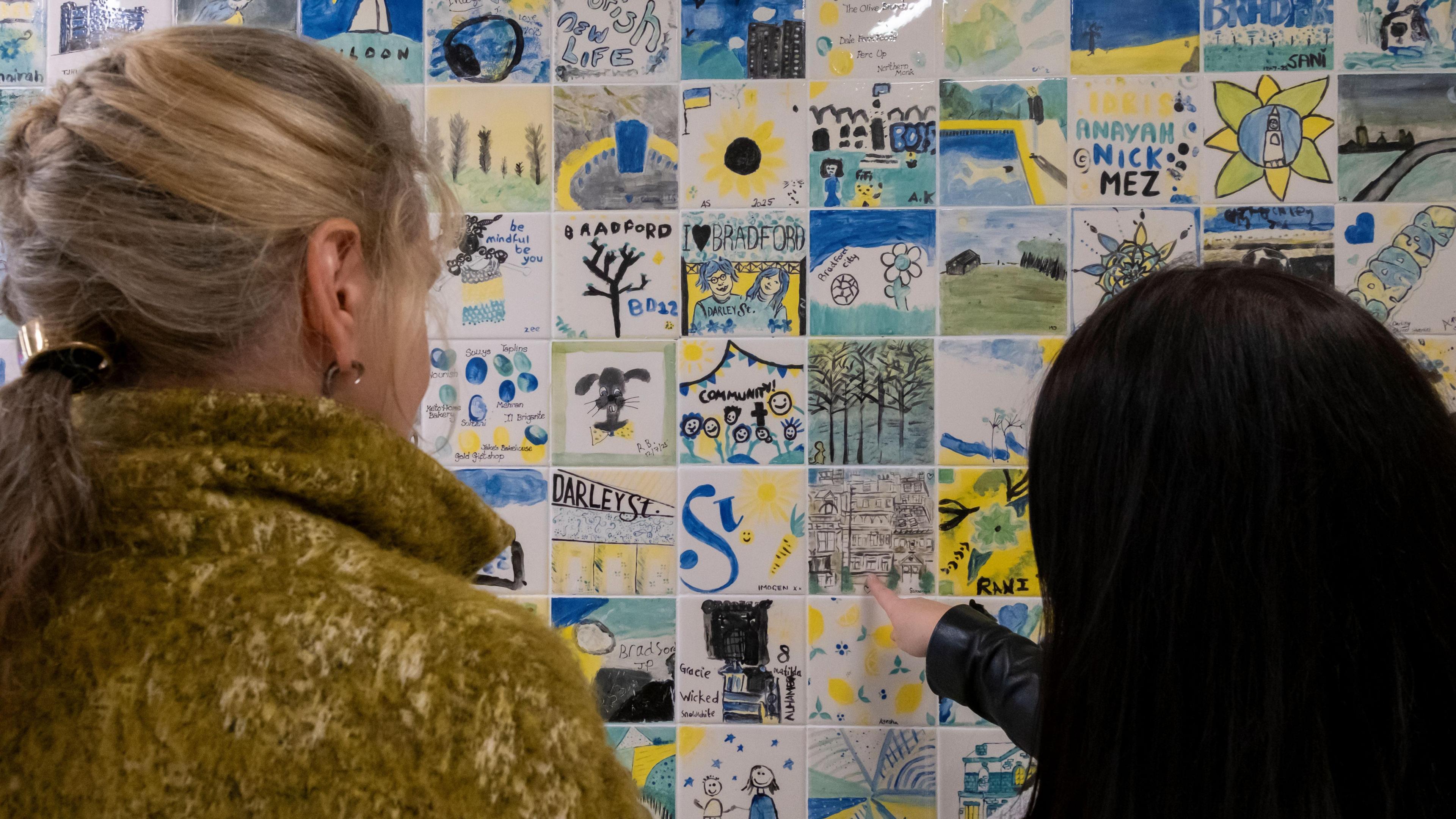 Two people are looking at a wall covered in small, colourful tiles with drawings and text.