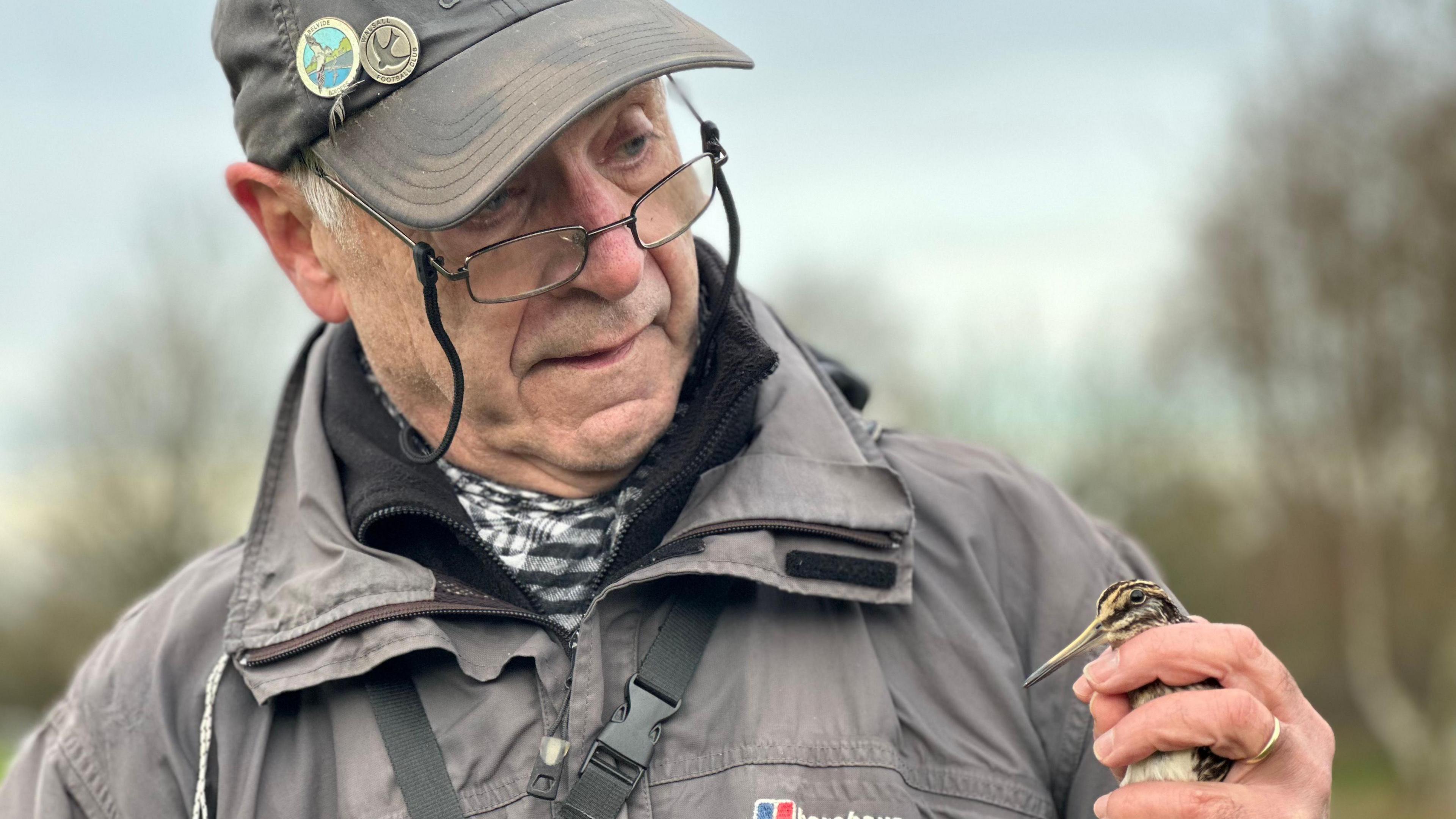 A man wearing a grey hat with two bird badges, glasses and a grey coat, looks at a jack snipe bird which he is holding in his left hand.