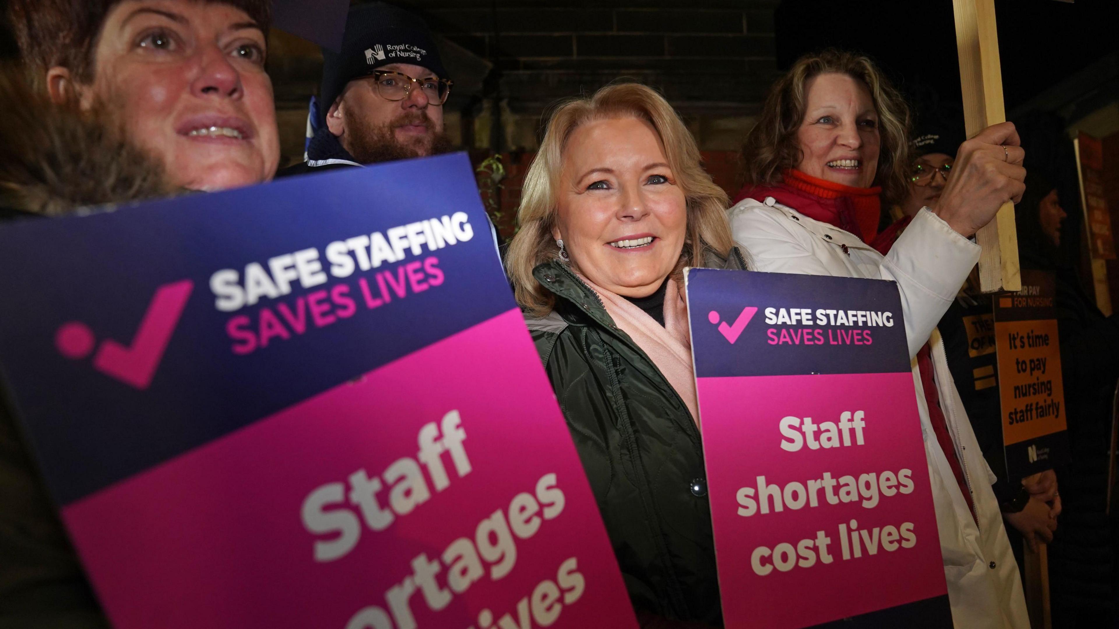 Pat Cullen, a woman with shoulder-length blonde hair, smiles as she joins a picket outside a hospital in the dark in Newcastle, England. There are two women stood either side of her holding purple and pink signs saying 'Safe staffing saves lives. Staff shortages cost lives' and one man with glasses standing behind her. 