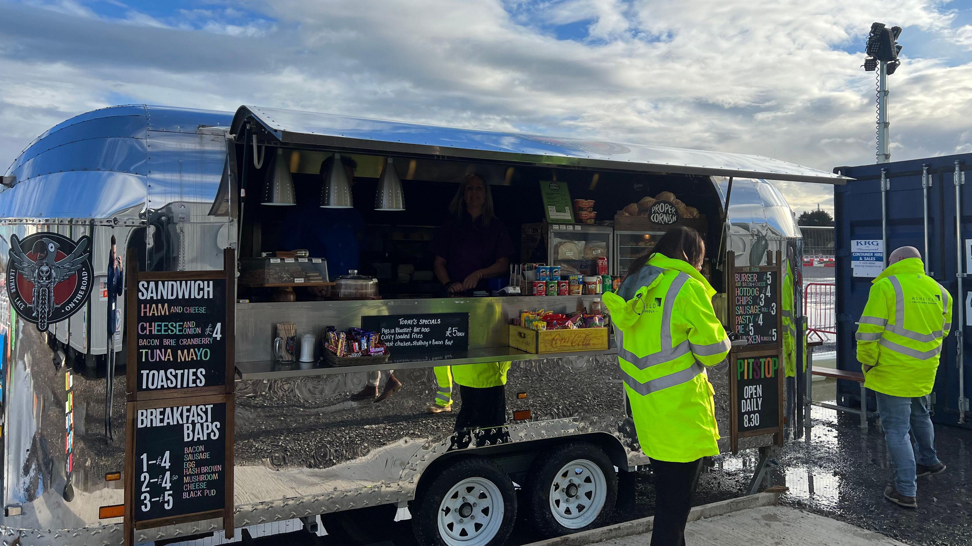 Big tin van shaped like a caravan. There's food and drinks on the counter with a bakery area and lots of crisps. There's two workers in high-viz jackets waiting to order