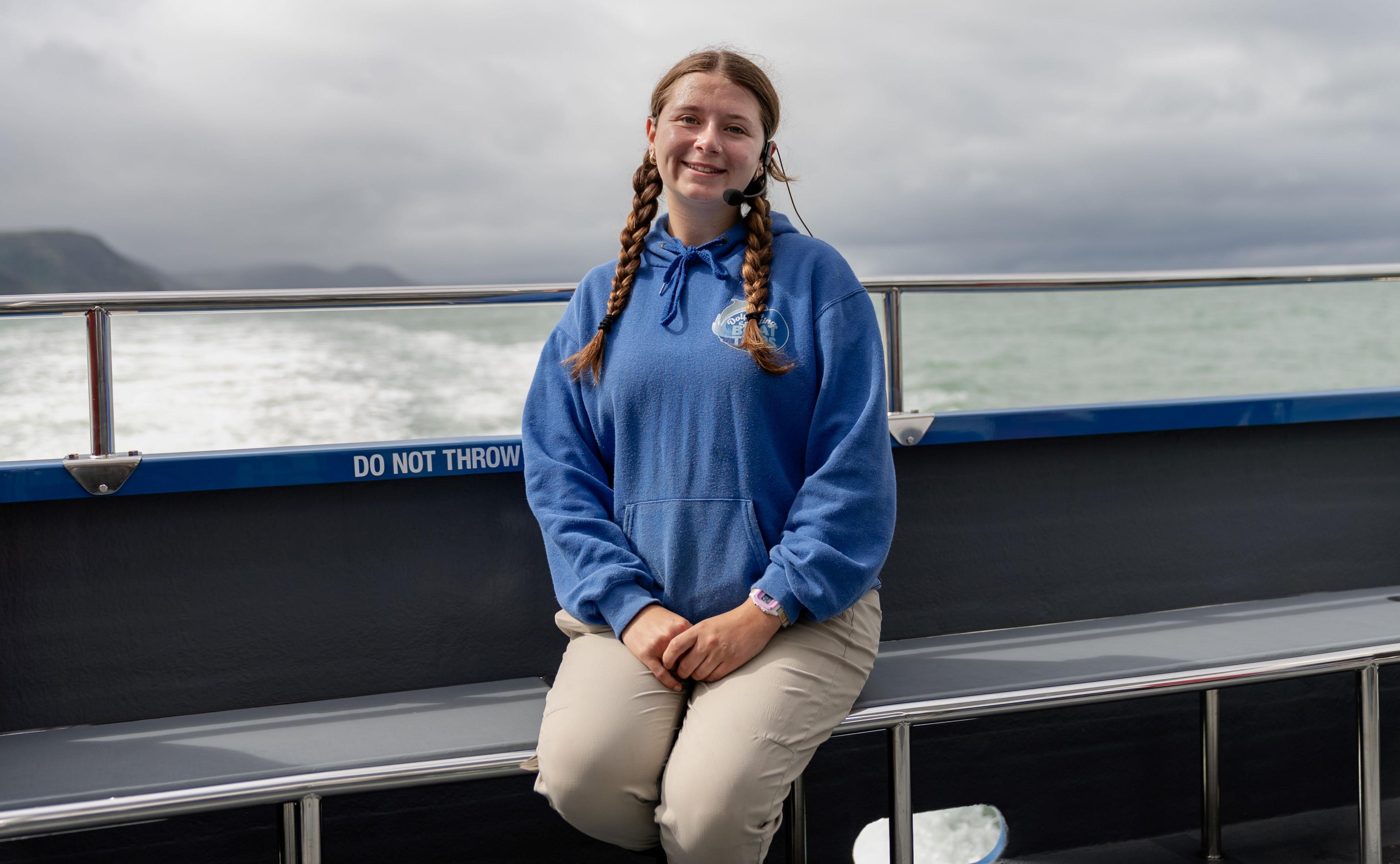 Suncatcher crew member Lauren Walthour who has her brown hair in two plaits, is hearing a mic head set and is sitting on the boat in a blue hoodie.