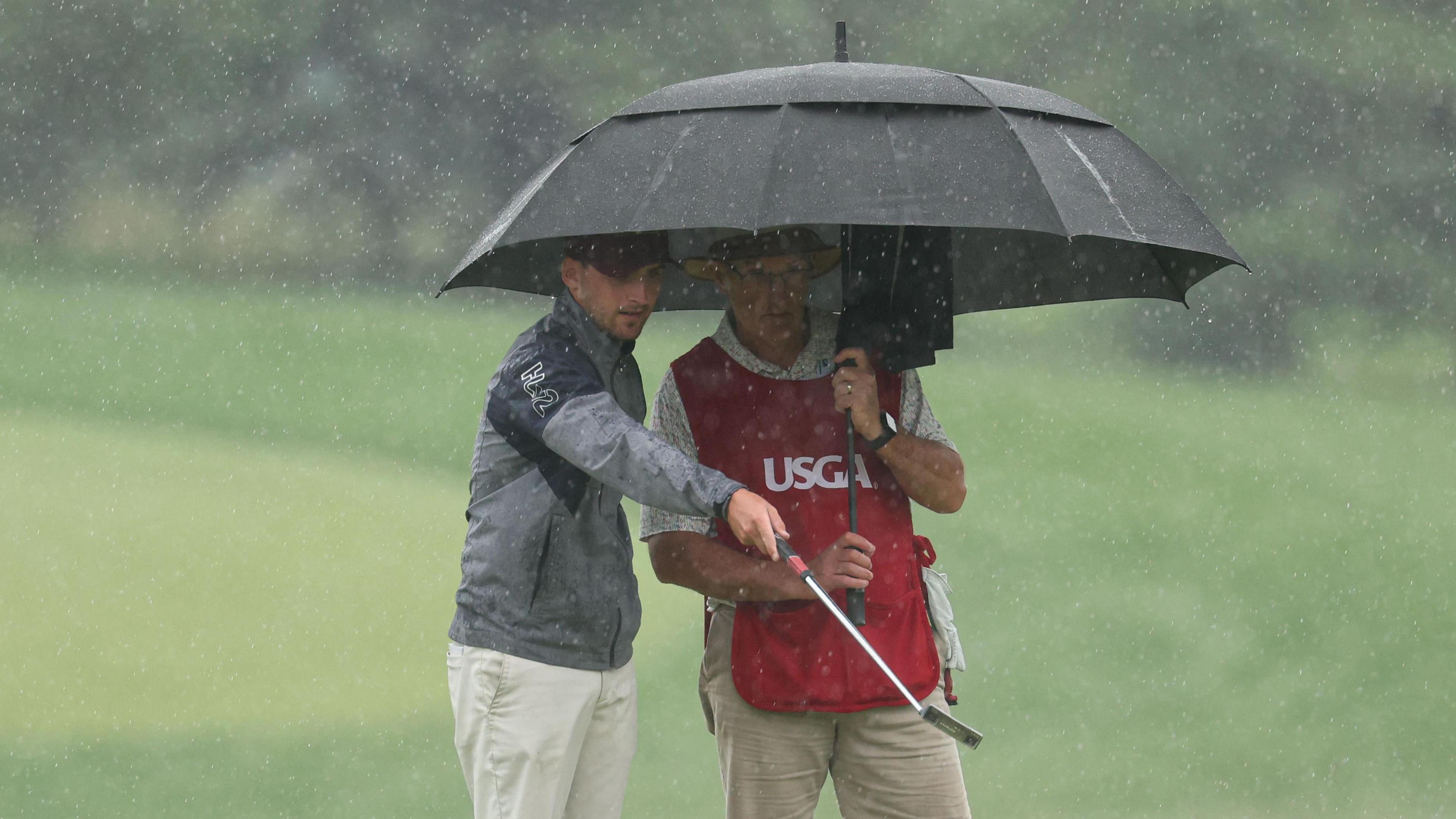 Jack Whaley pointing at the ground with his golf club while he and Andy Whaley, his father and caddy, stand under an umbrella with rain visible in the photograph