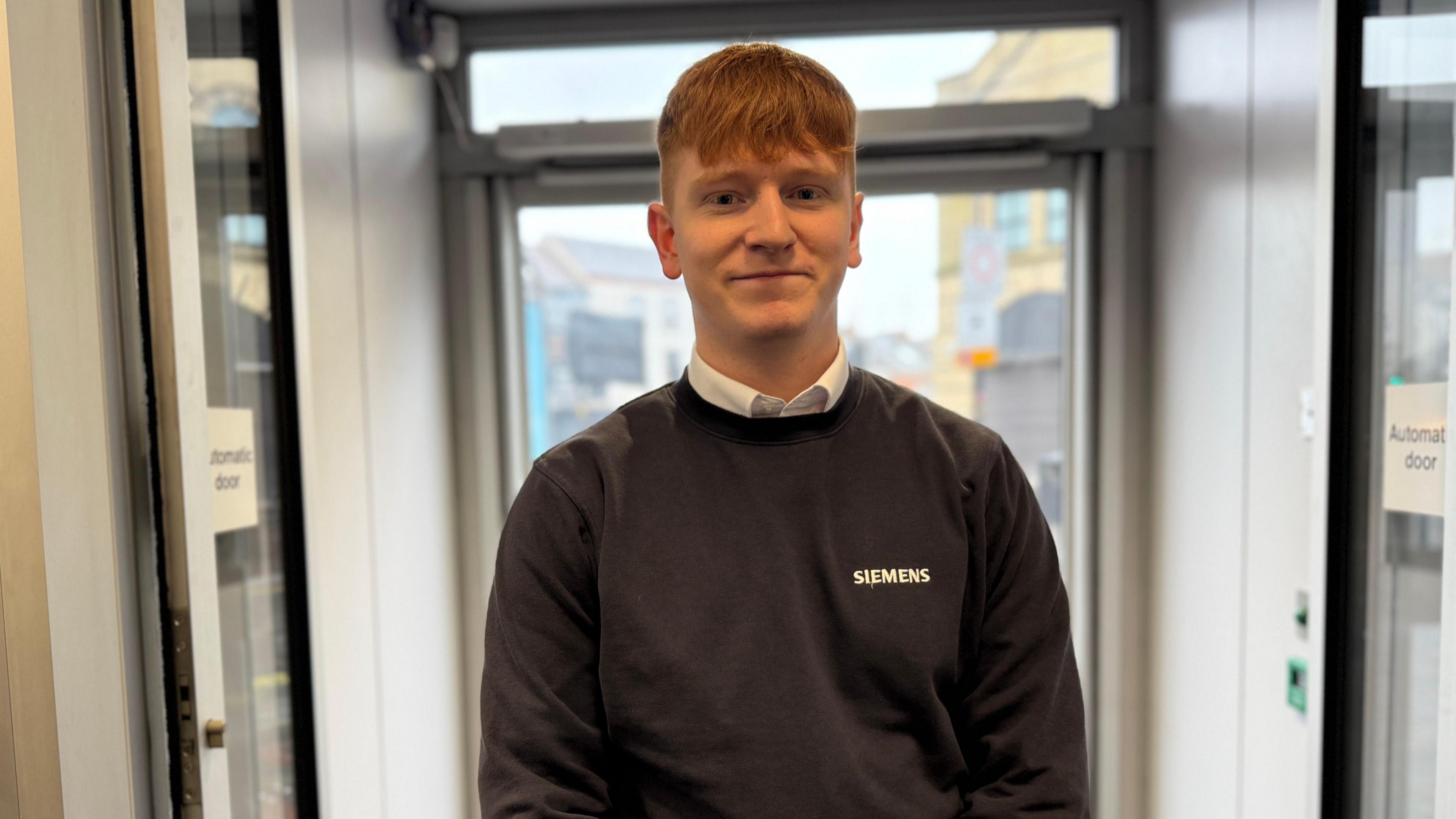 A man with ginger hair in a grey jumper and white shirt smiles. 