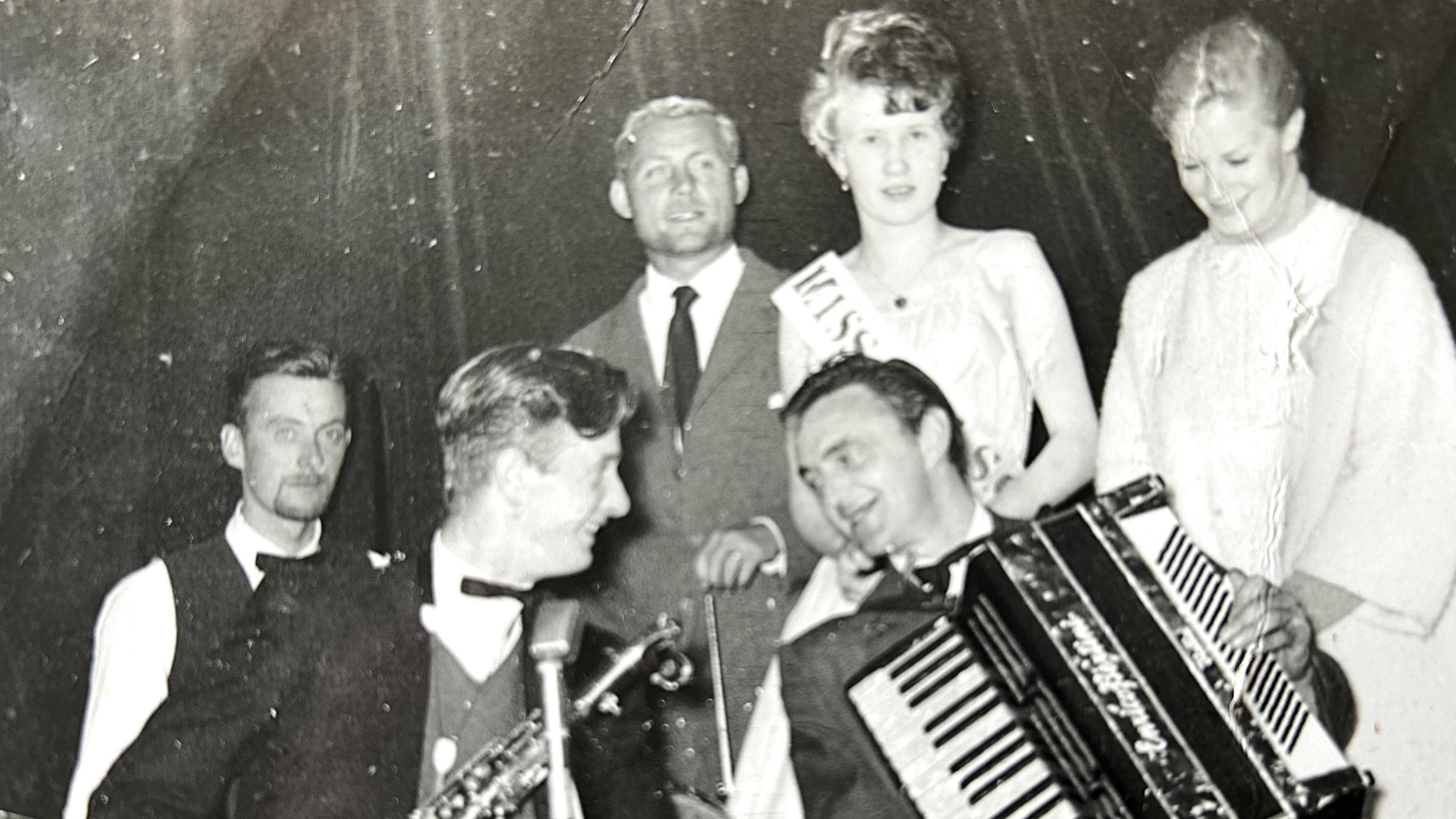 Six people standing together. One has an accordion and his bandmate has a saxophone. A third band member sits to the left of them but he has no instrument. Behind them stands Robert Shaw in a grey suit, white shirt, dark tie. A female winner in a light dress and a winner's sash, and Mary Ure in a white coat. They all appear to be enjoying themselves.