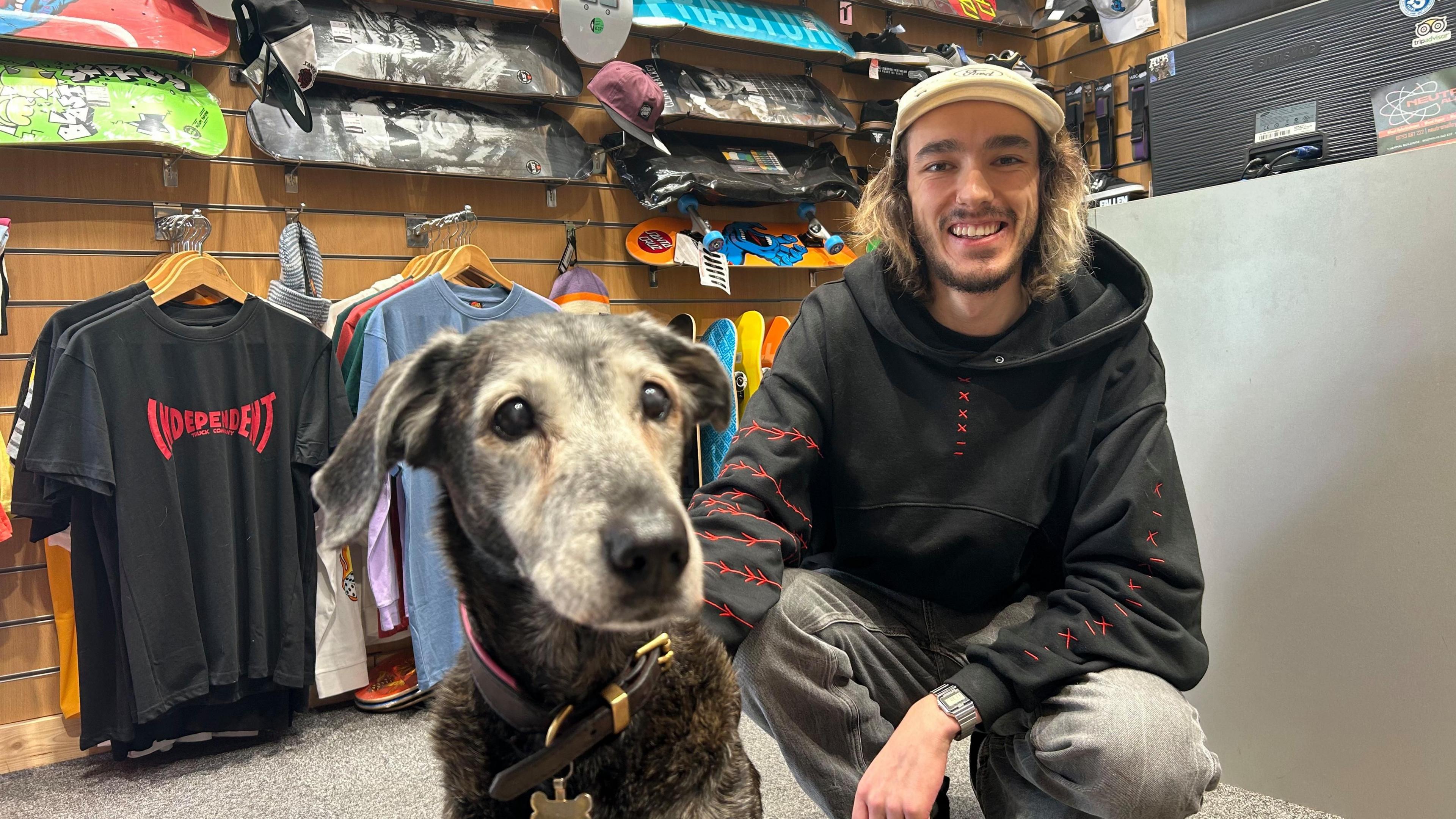 Abbie sits on the floor with Tommy who is crouching. He wears a black hoodie, a white hat and grey jeans. Behind them is the shops front desk and to the left are rails of T-shirts. 