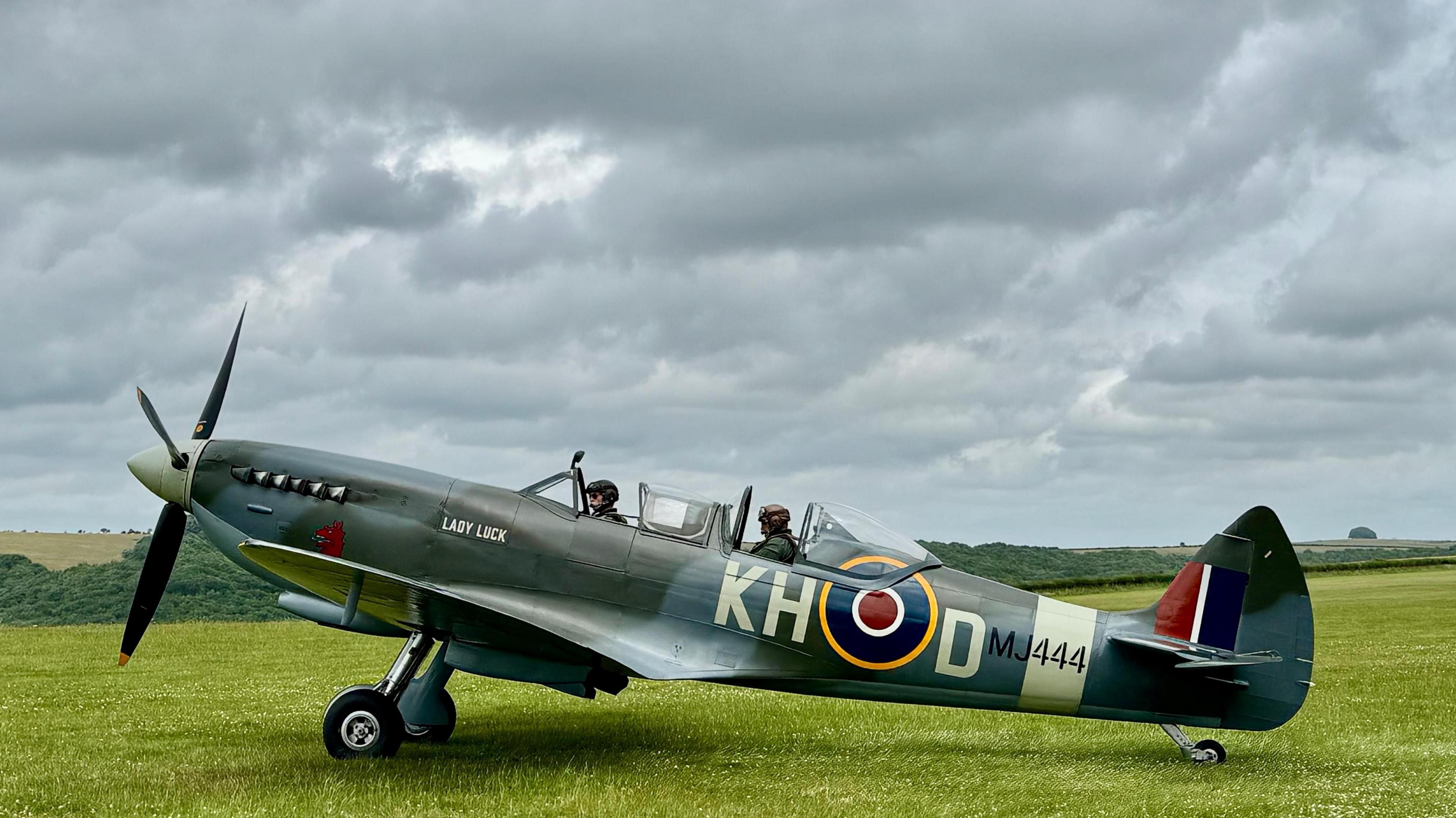 A spitfire sits on the grass, preparing to take off with two people on board. The skies above are grey and full of clouds