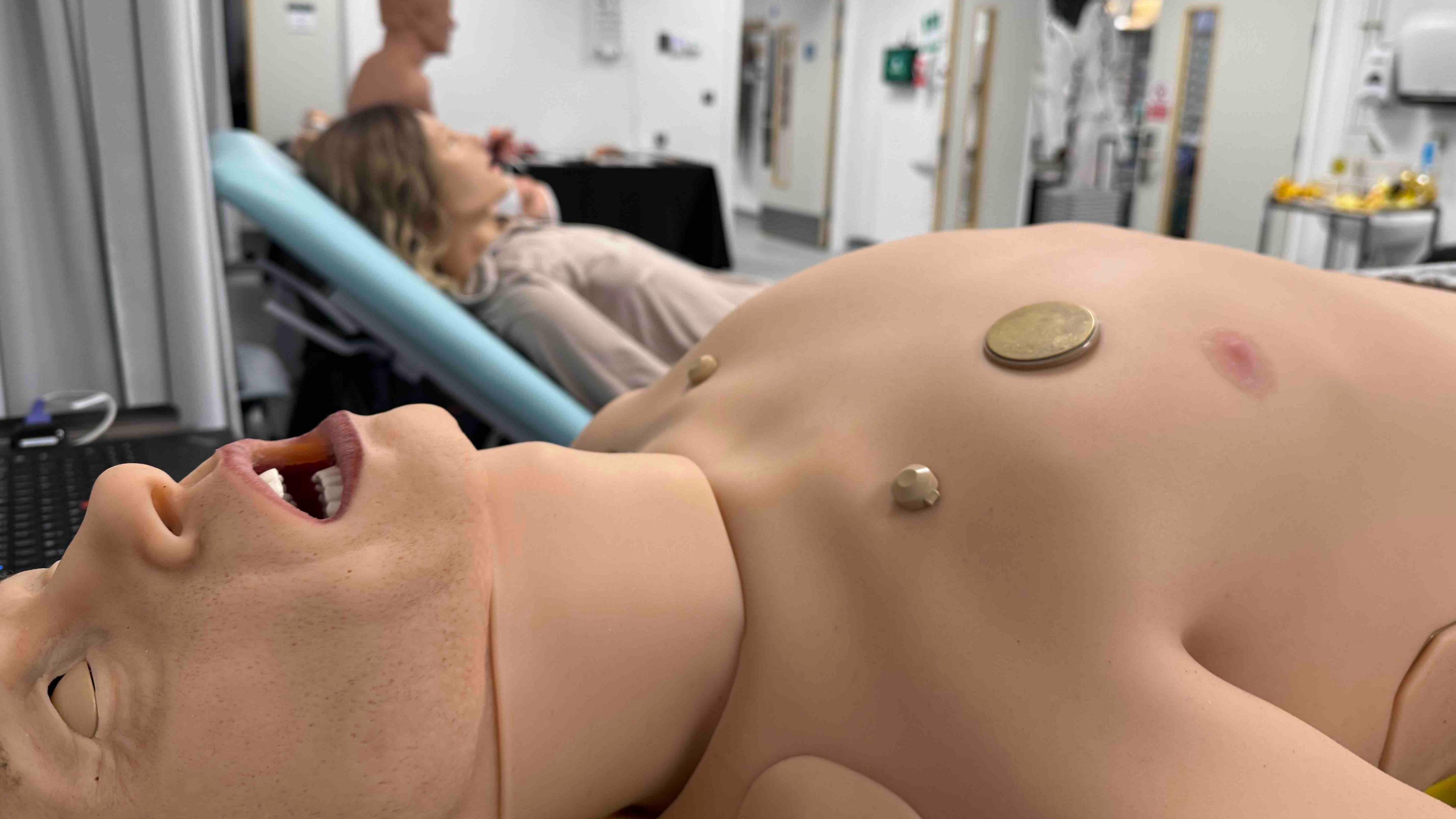 A close-up of a lifelike mannequin lying on a medical bed in a large room resembling a hospital ward. The mannequin has an open mouth, closed eyelids and three round vents, with gold-coloured covers, on its chest. More mannequins can bee seen on beds in the background, including one with long hair and another sitting up.