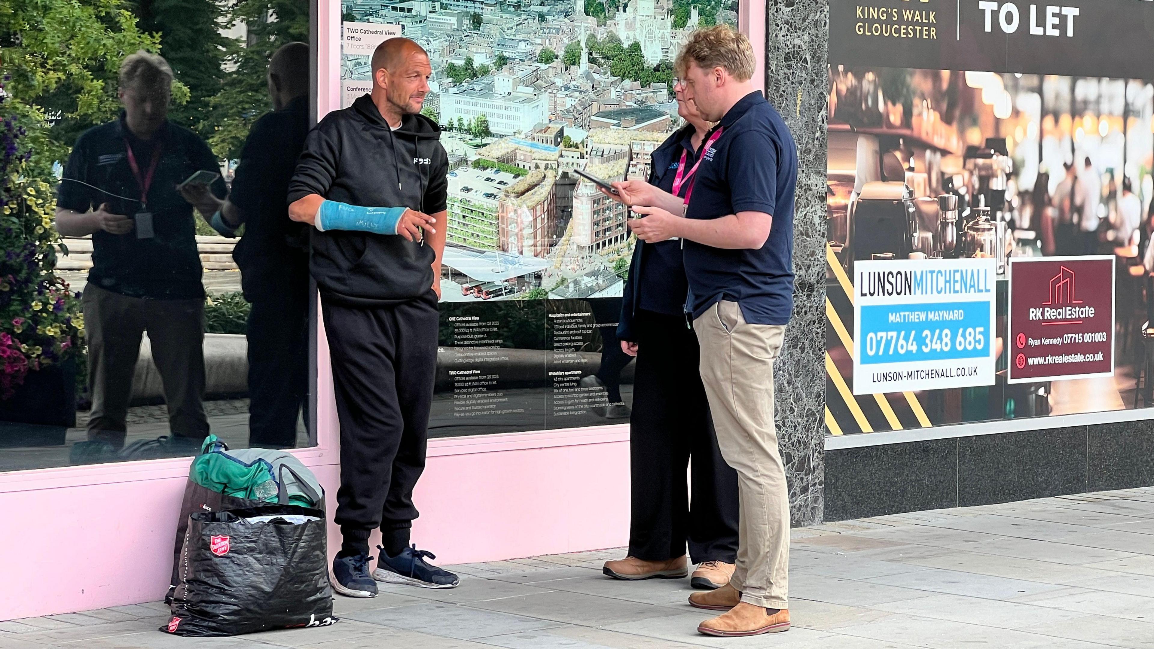 A man, who is bald and wearing a blue arm cast and black clothing, stands outside a shop while two council officers speak to him. There are a couple of shopping bags containing sleeping bags beside him.