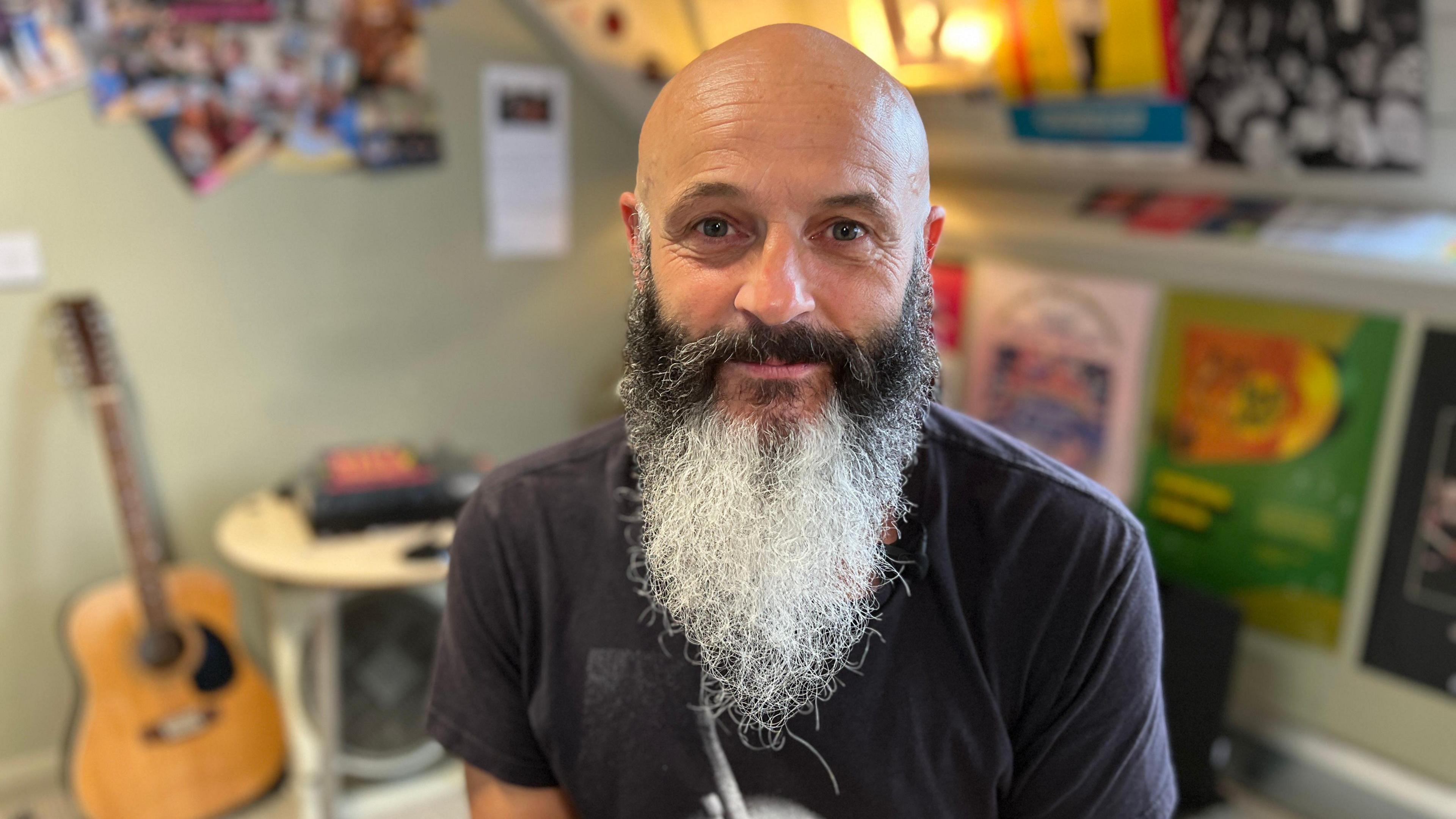 A man with a long, greying beard sat in a room. There is a guitar and a desk behind him, along with a wall covered in posters. He is smiling.