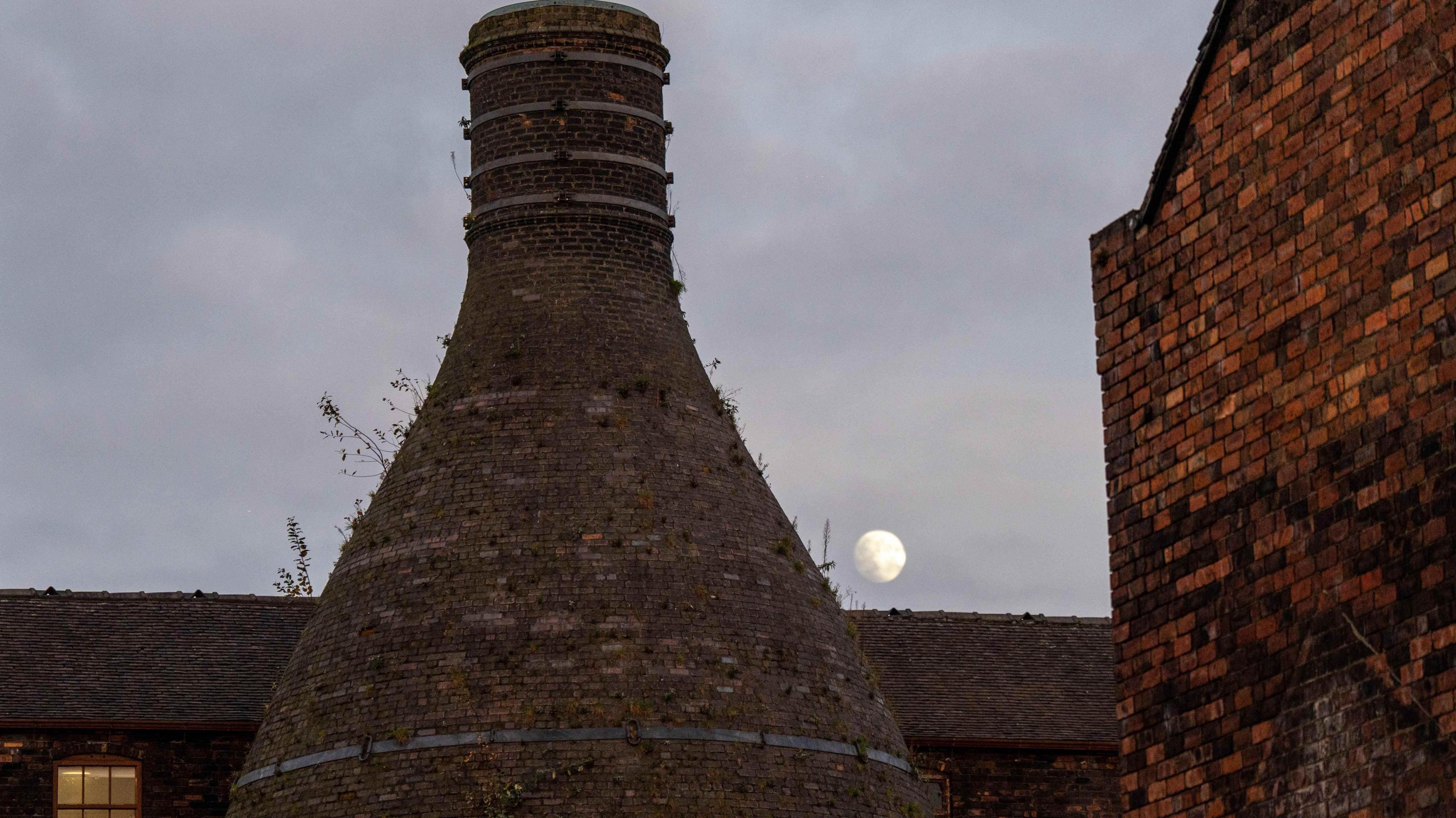 A bottle oven and brick buildings in the foreground with a full moon visible in the sky in the background.