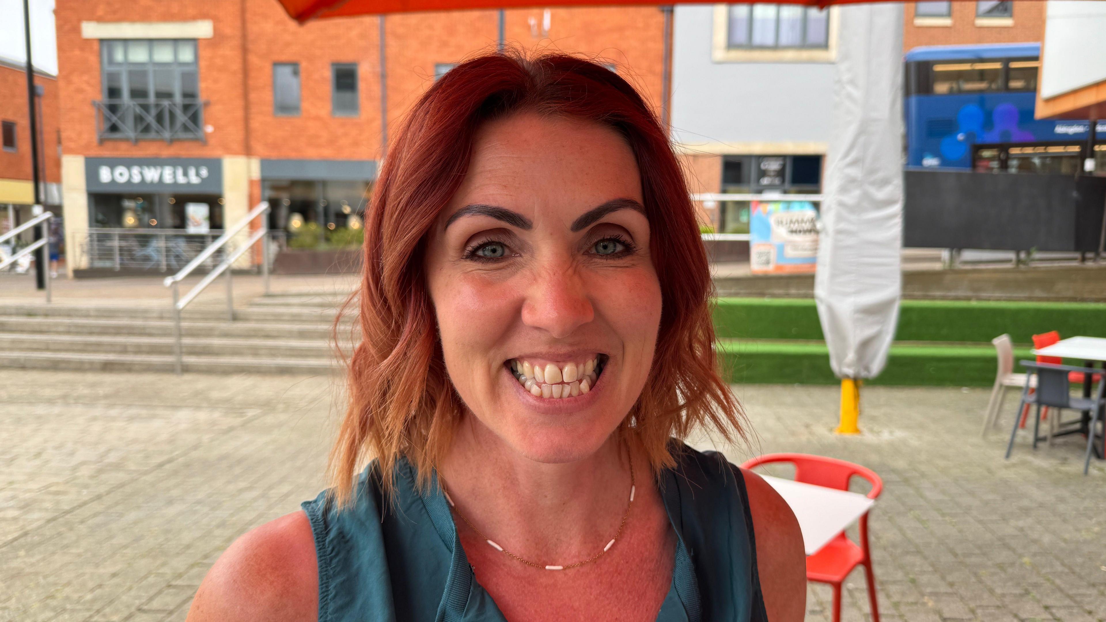Ruth Davies smiles for the camera. She is around 40-50 years old with neck-length dyed red hair. She's wearing a small necklace and a turquoise top. Cafe tables and chairs can be seen in the background