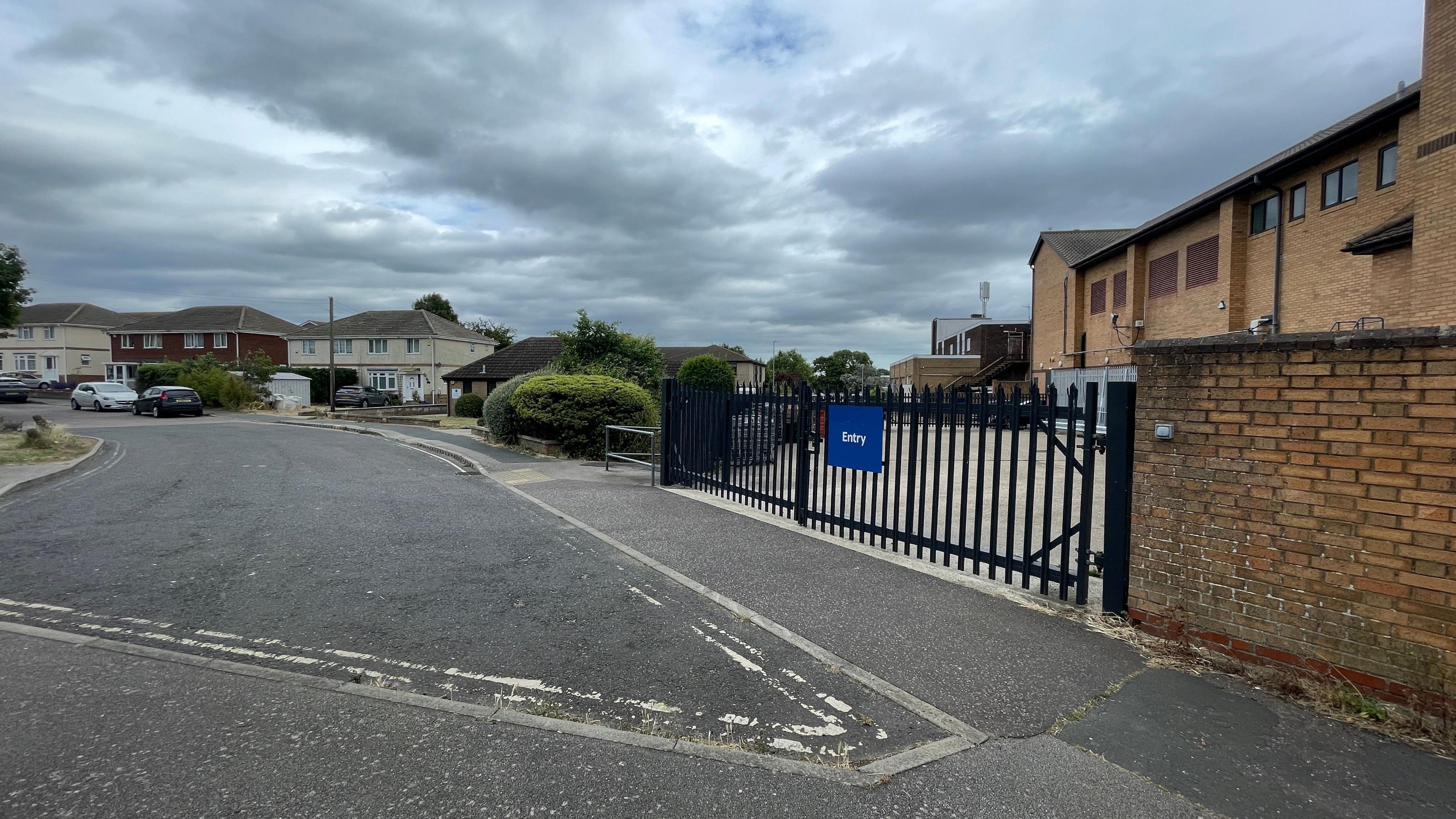An area in Walton-on-the-Naze in Essex where an attack on an 82-year-old man took place. The area has a road and brown bricked buildings. There is also a black fence with a blue rectangular sign on it which says Entry. 