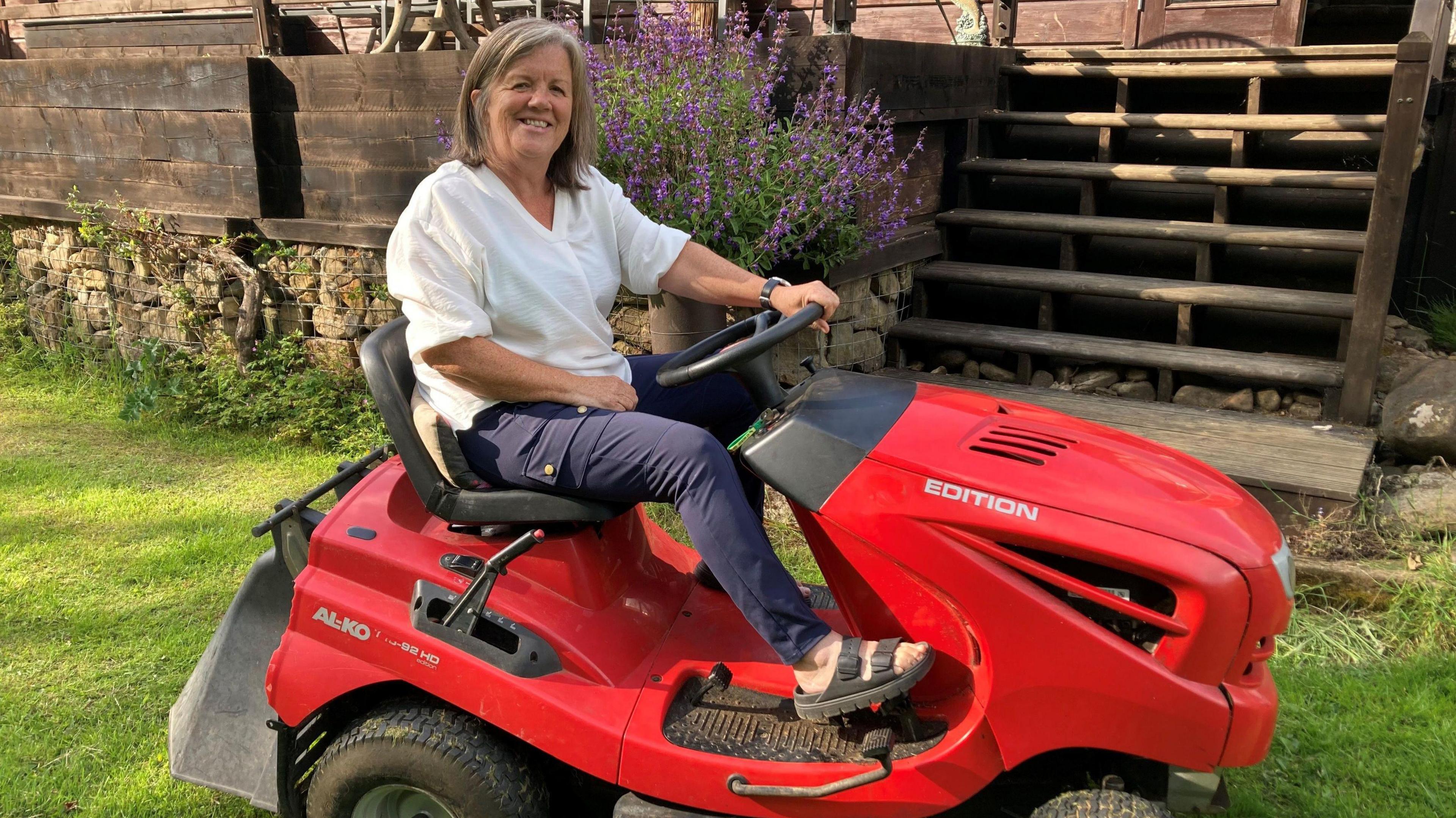Catherine is sitting on a red tractor mower. She is wearing a white top and blue trousers. She is in front of a wooden platform on grass.