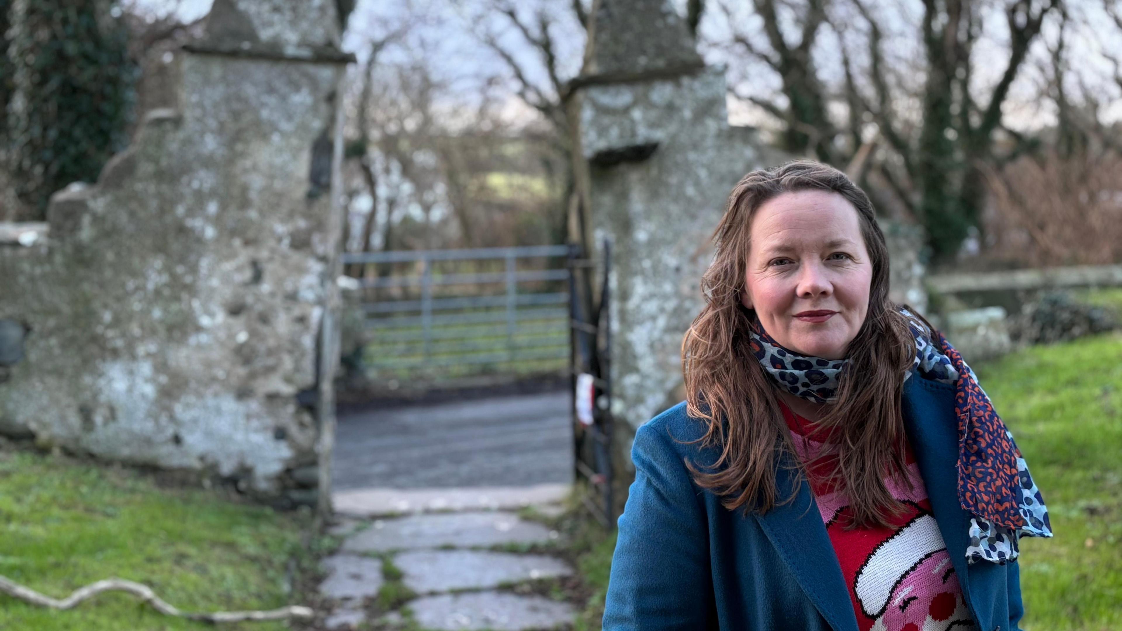 Caroline Devlin, a woman with brown hair, wearing a blue blazer and a red Christmas jumper and a scarf, with the slanted gate posts of the church in the background.