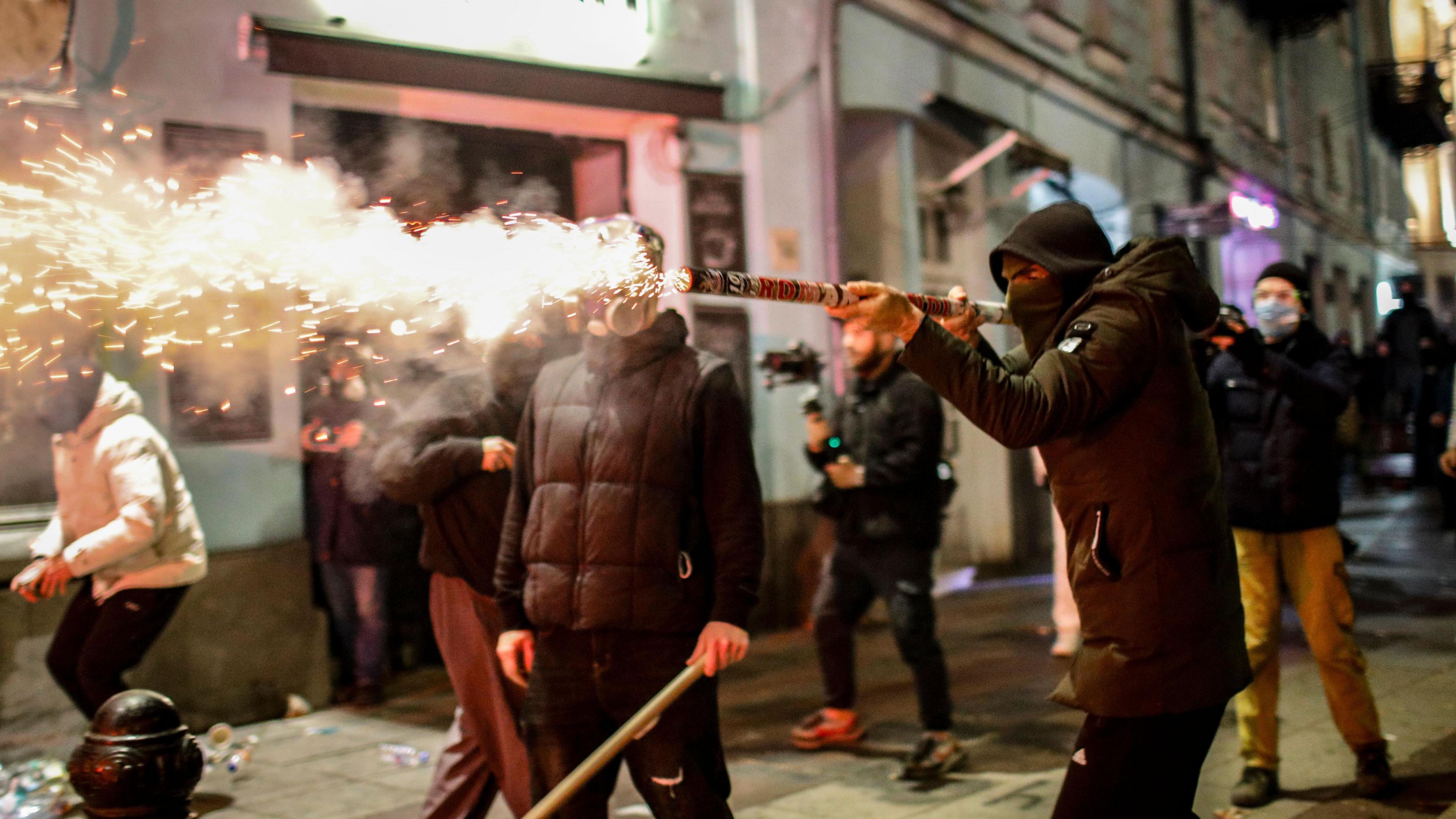 A Georgian opposition supporter in a jacket launches a firework towards police. He is wearing a hood and a mask and is surrounded by other protesters