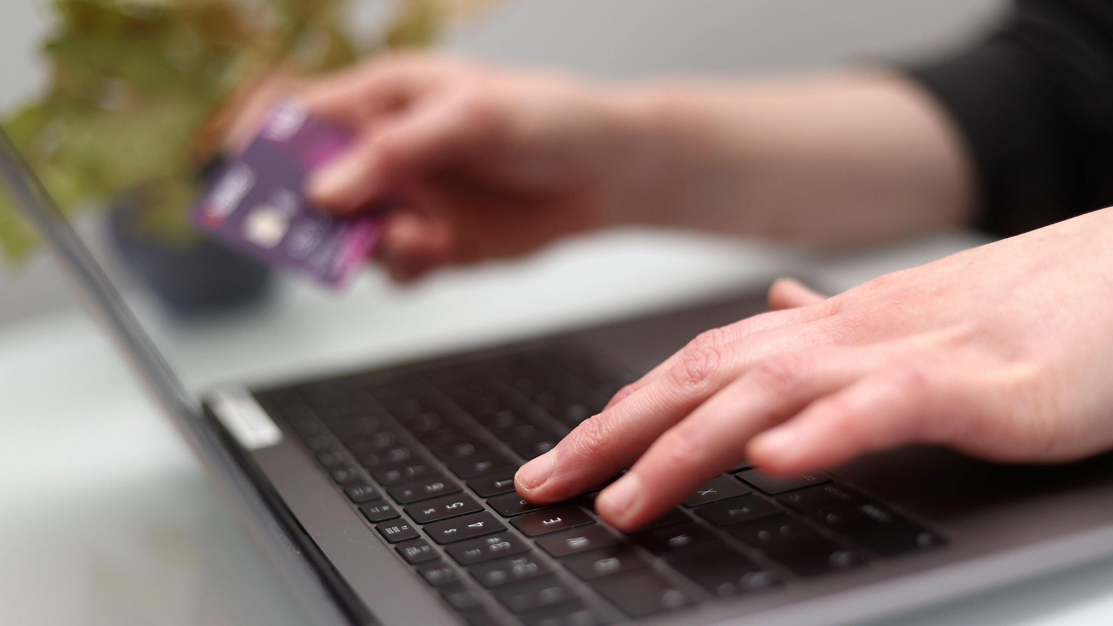 A woman holds a credit card in her right hand while typing with her right hand on a grey laptop.