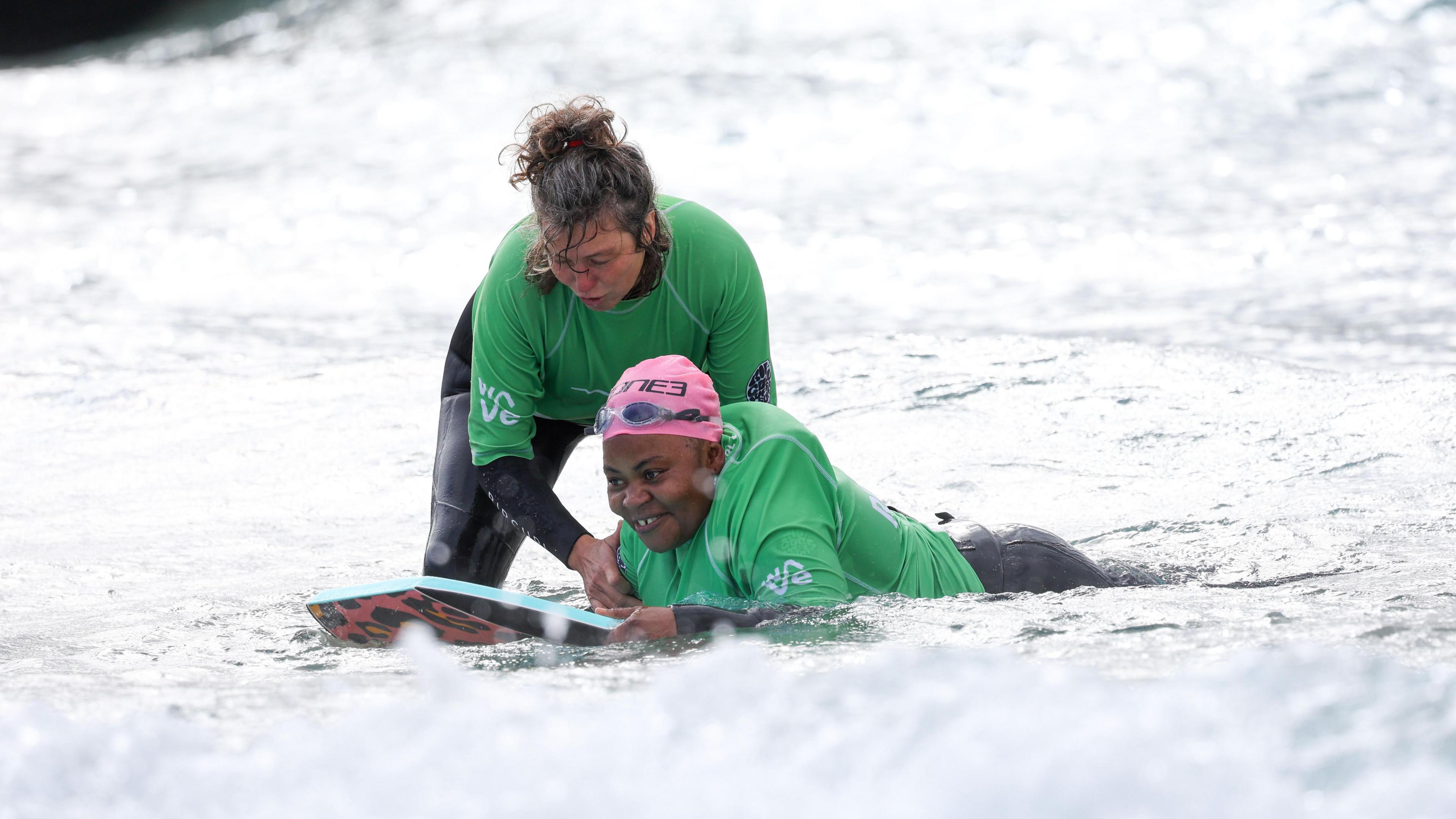 A woman lying on a bodyboard at The Wave in Bristol. She is wearing a wetsuit, green rash vest and pink swimming cap. A female volunteer is standing beside her and holding her shoulder, ready to guide her into catching a wave.