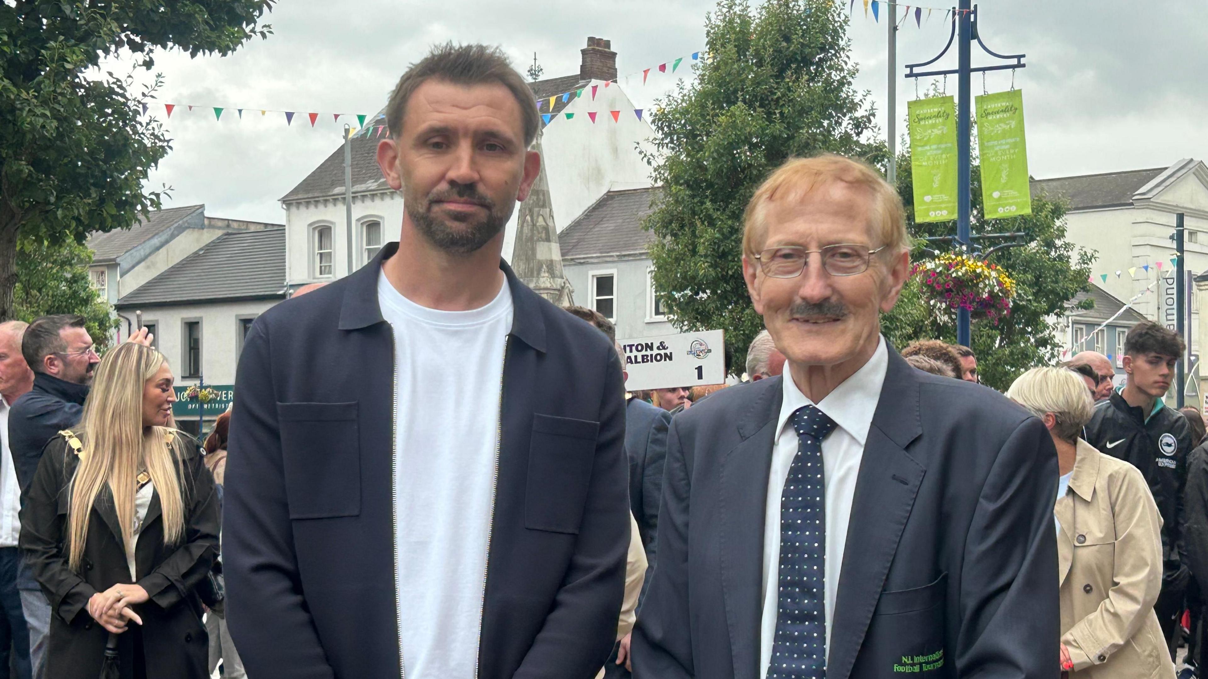 Two men pose for a photo. One is wearing a white top and a navy jacket and one is wearing a suit. Crowds are behind them. 
