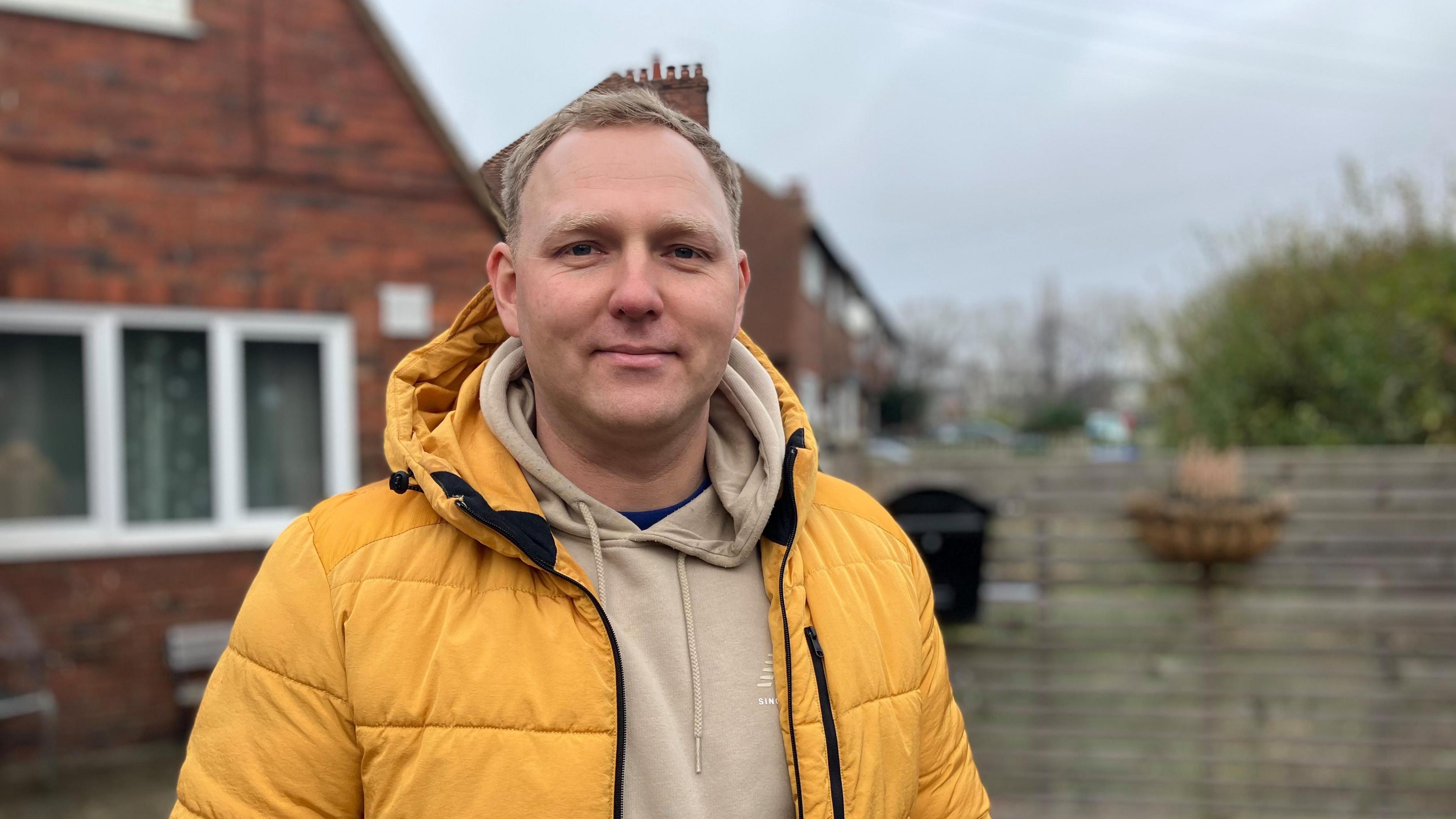 Karolis Arlauskas is seen looking forward standing outside his property. He is wearing a hoodie with yellow coat. His red bricked home and fence can be seein the background.