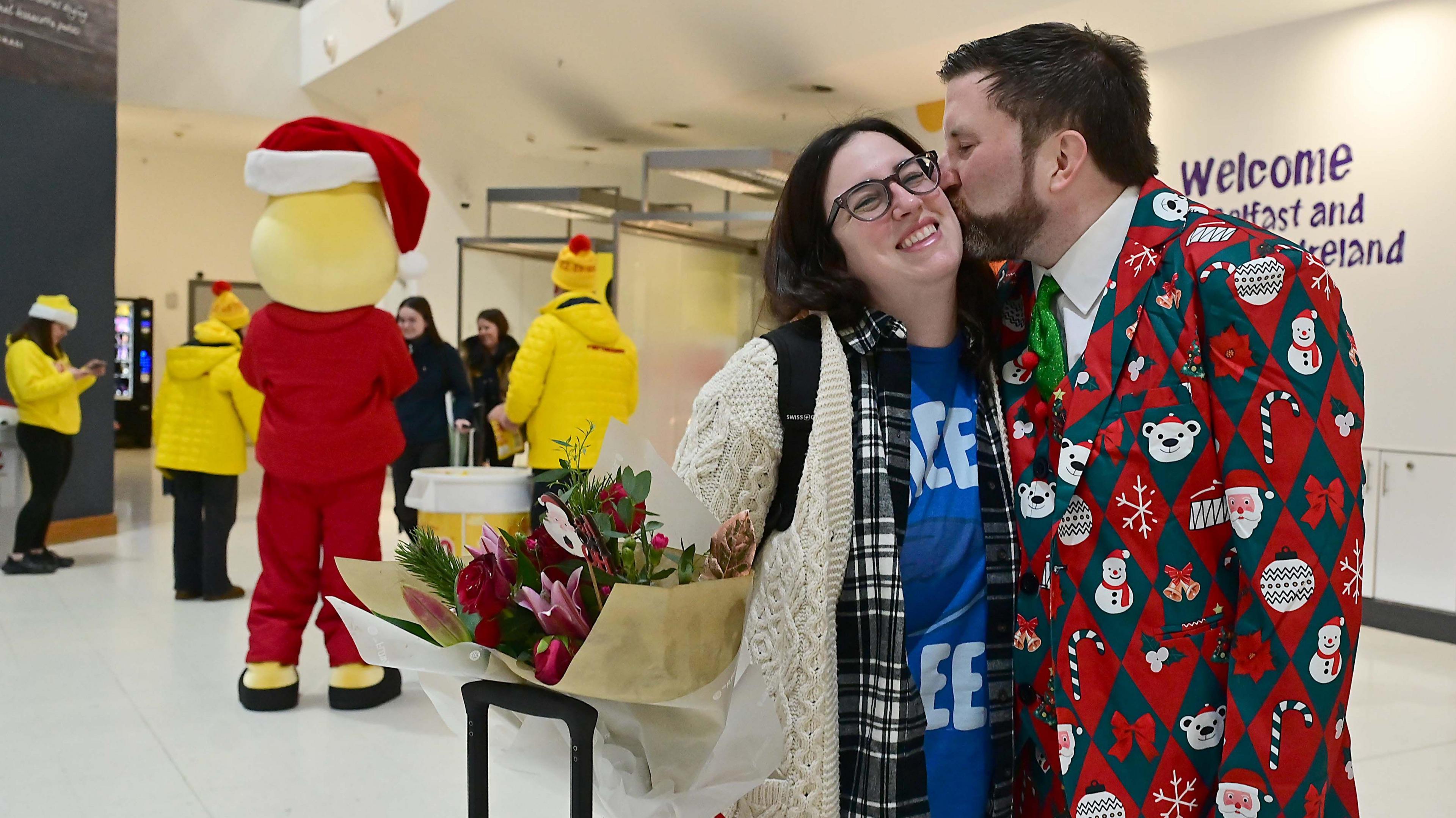 A woman with brown hair, a cream cardigan and a bunch of flowers is kissed on the cheek by a man in a Christmas themed suit - which is red and green, and features snowmen, polarbears, snowflakes and candy canes. He has brown hair and a beard. 