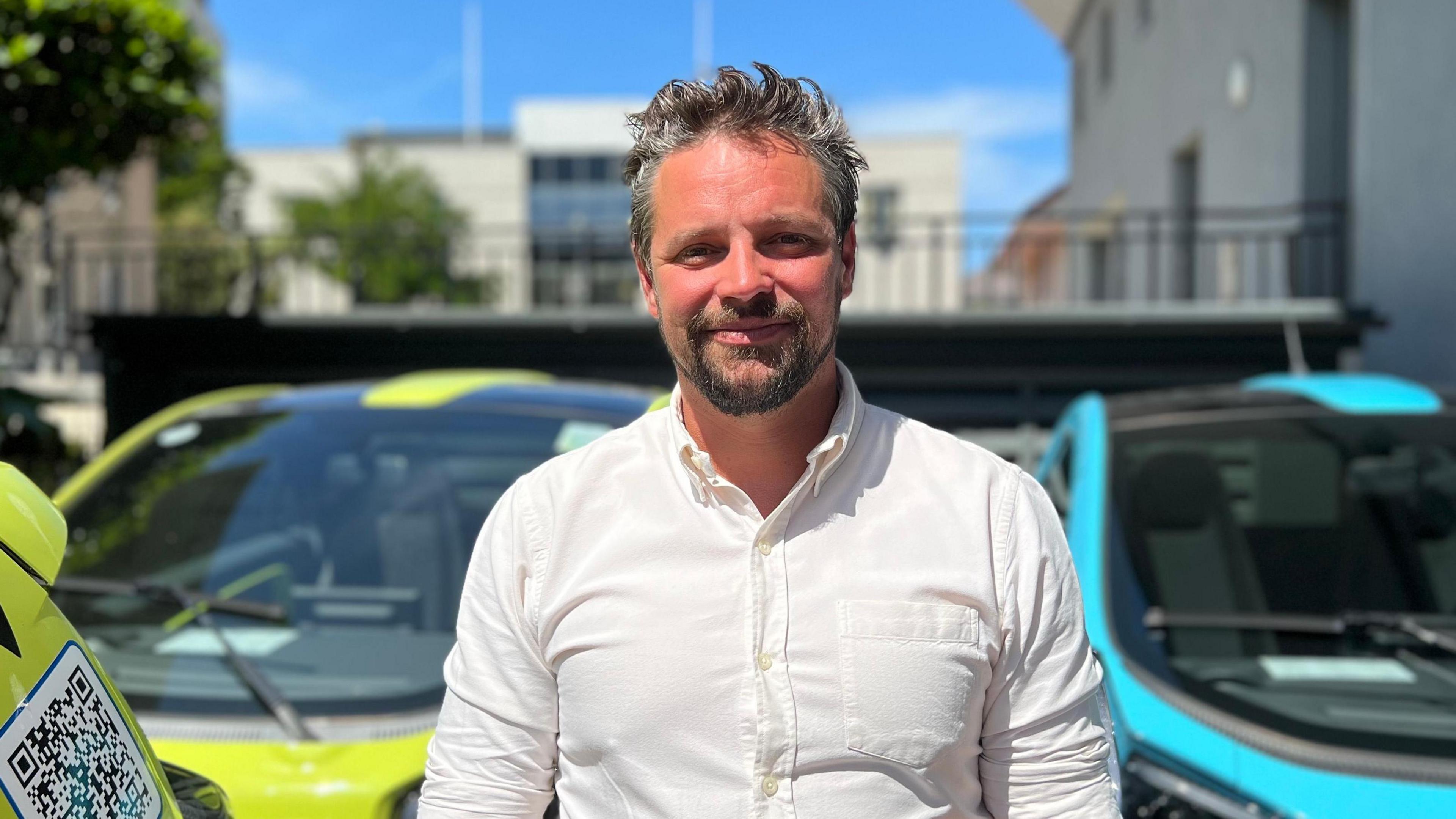 Nick Perchard-Rees is a man standing in the sunshine, smiling into the camera. He is standing in front of a green and a blue EVie hire car.