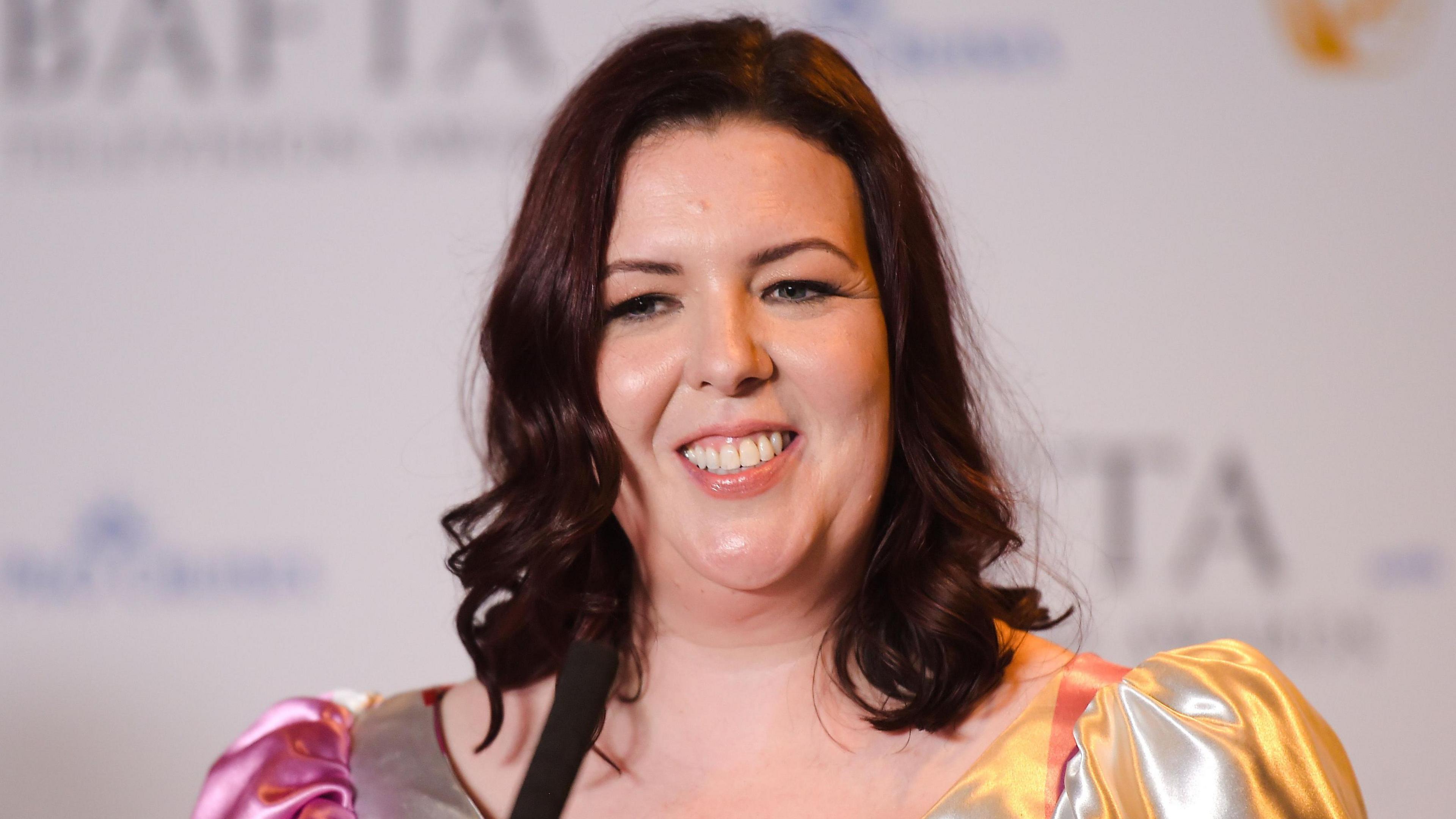 Lisa McGee smiling on the red carpet at the Bafta TV awards 2023. She has shoulder-length brown curly hair and is wearing a silver dress.