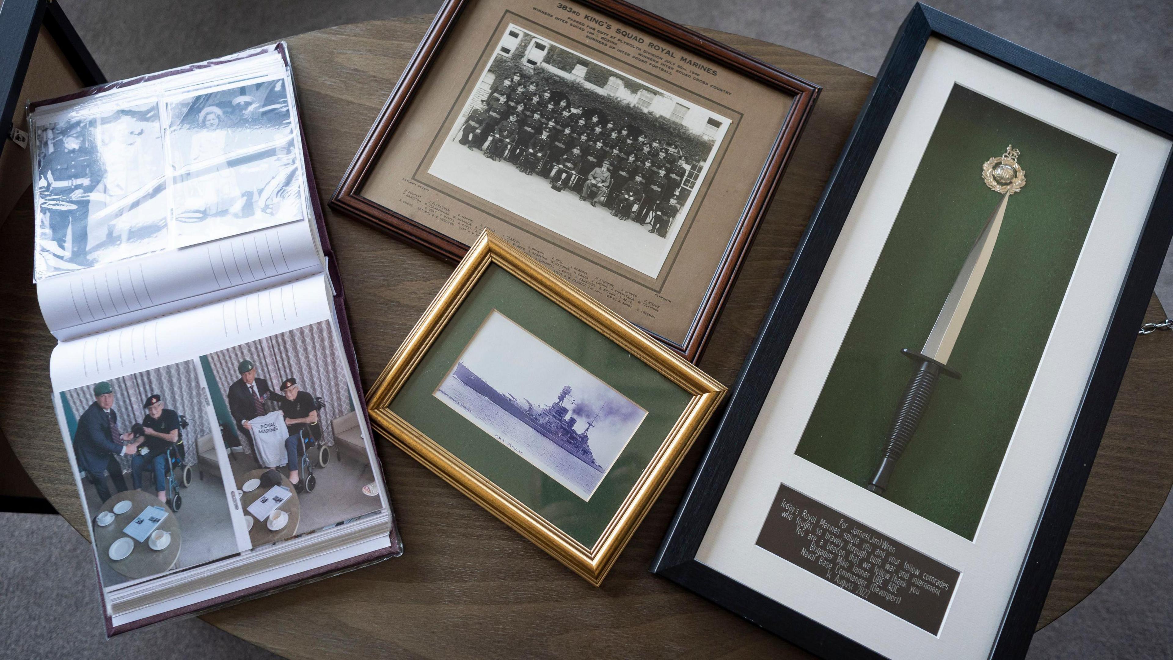 A photo album, old framed photos and a framed commando dagger on a table. The photos depict people and a large group of Royal Marines who served in World War 2.