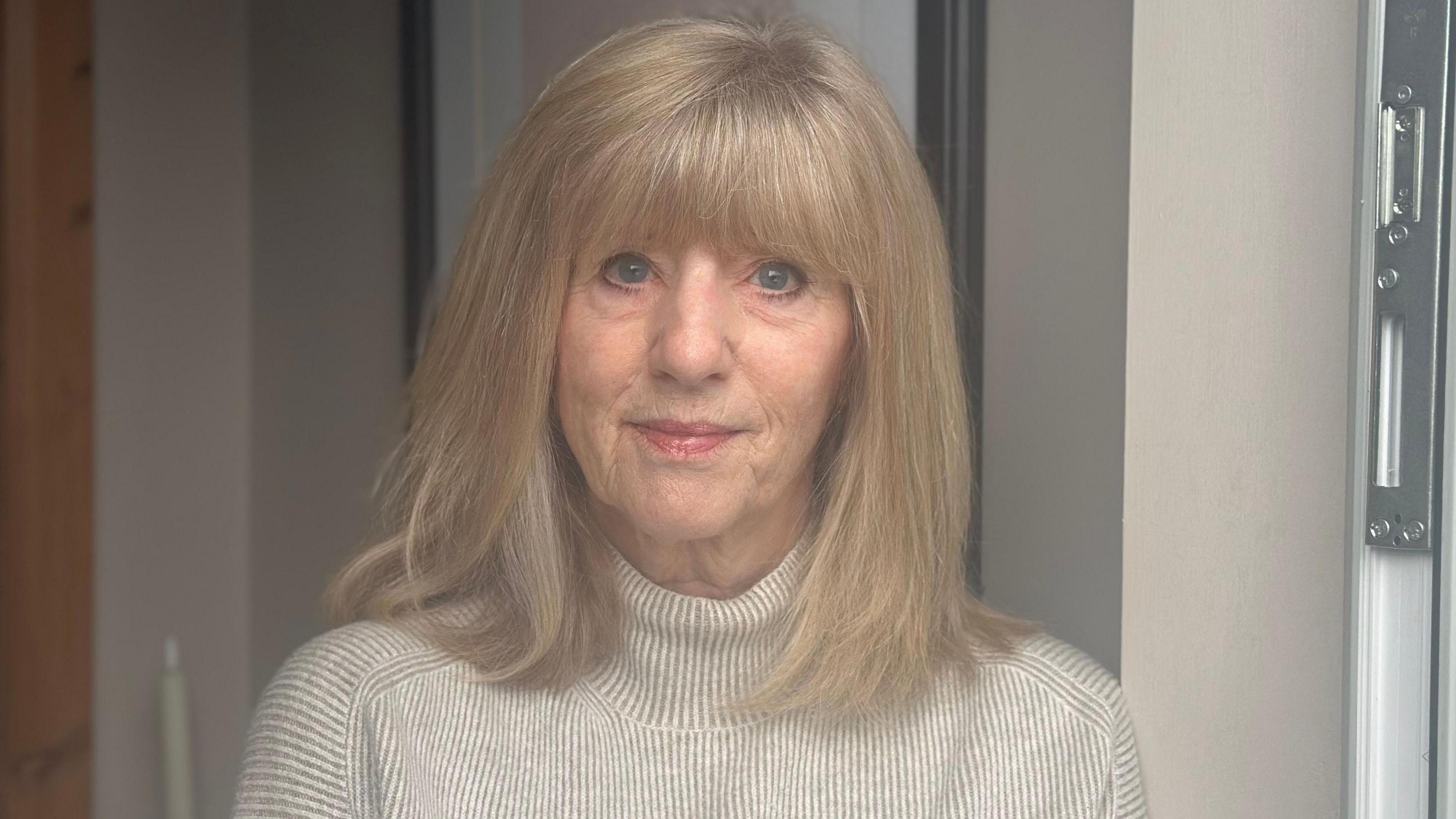 Janet Preston with long blonde hair and a beige polo neck standing at the front door of her house. There is a photoframe on the wall behind her.