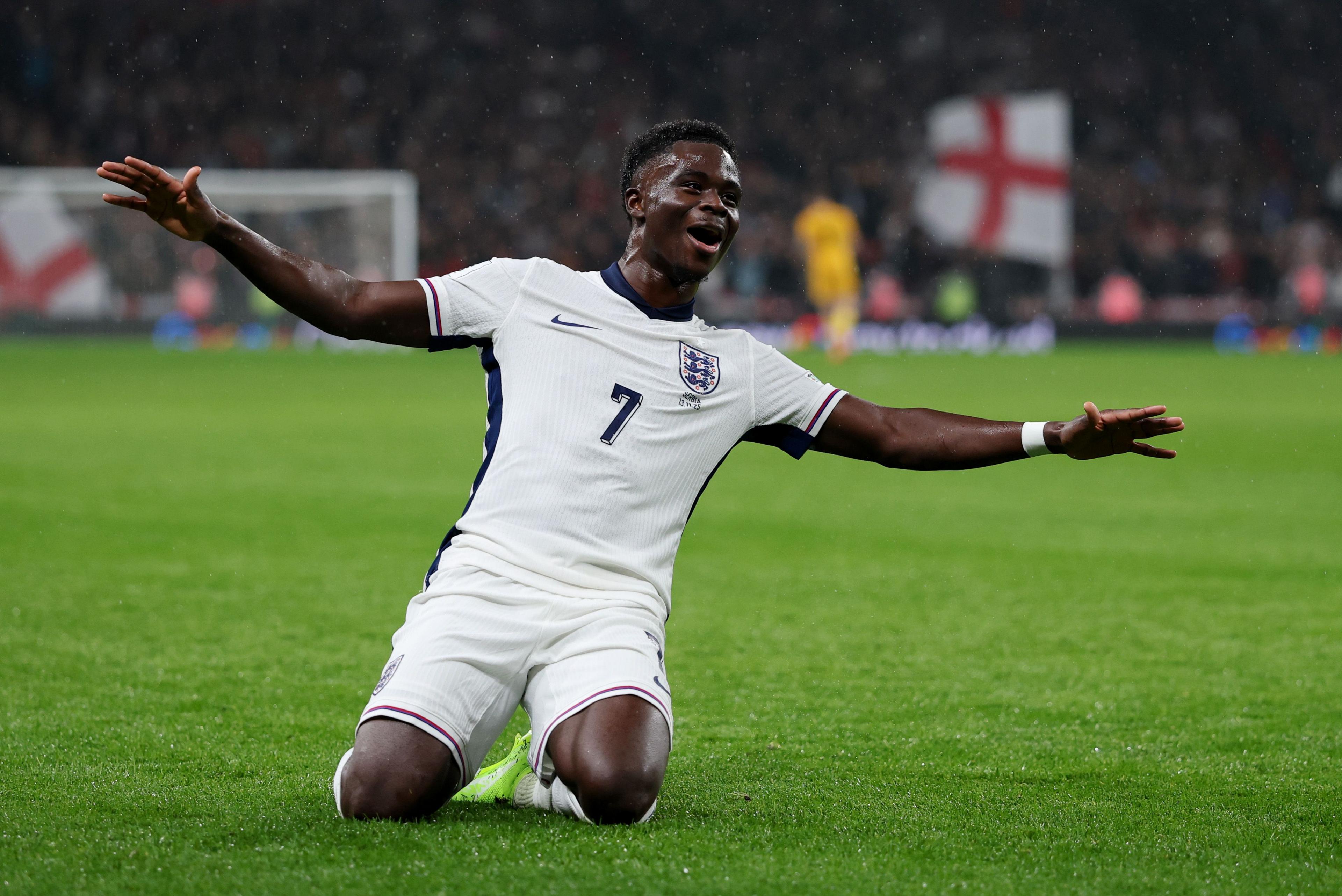 Football player in a white England kit sliding on knees with arms outstretched in celebration on a green pitch, with an England flag visible in the background.