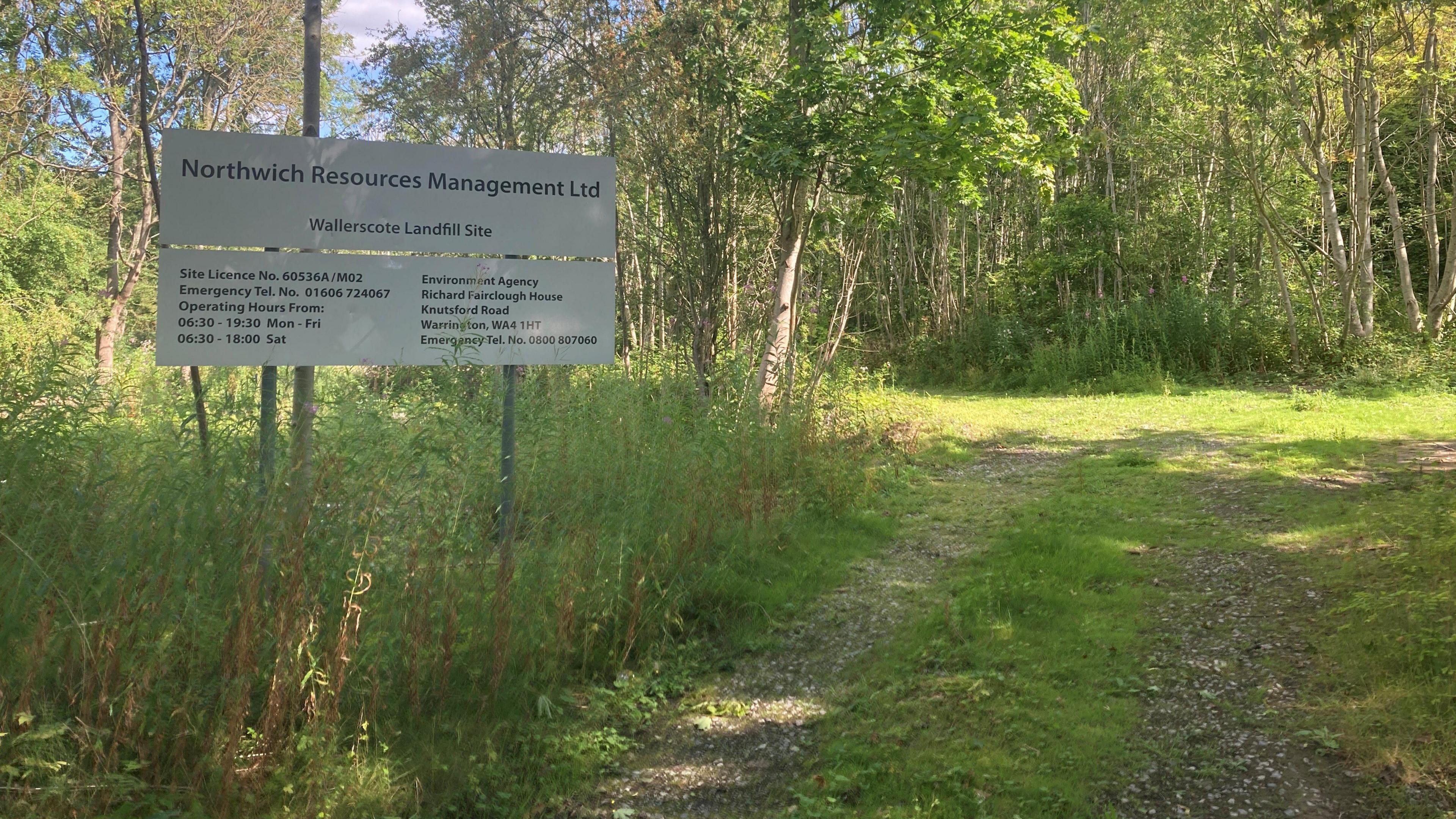 View of a sign for Wallerscote Landfill site. It has Northwich Resources Management written on the top, then Wallerscote Landfill Site beneath. There are also phone numbers and contact details on the site, which stands in overgrowth at the side of a gravel path.