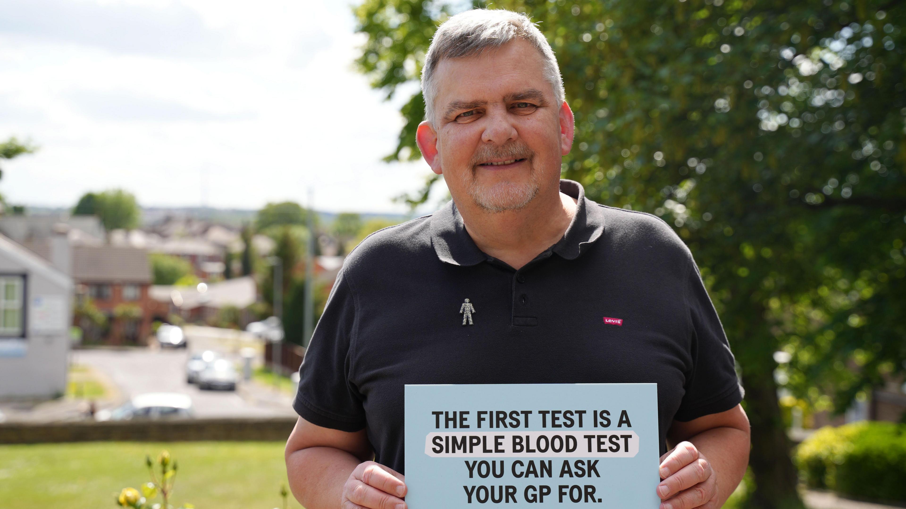 A man with grey hair and a beard stands in a garden. He wears a black polo shirt and a Prostate Cancer UK badge. He holds a card which reads: "The first test is a simple blood test you can ask your GP for."