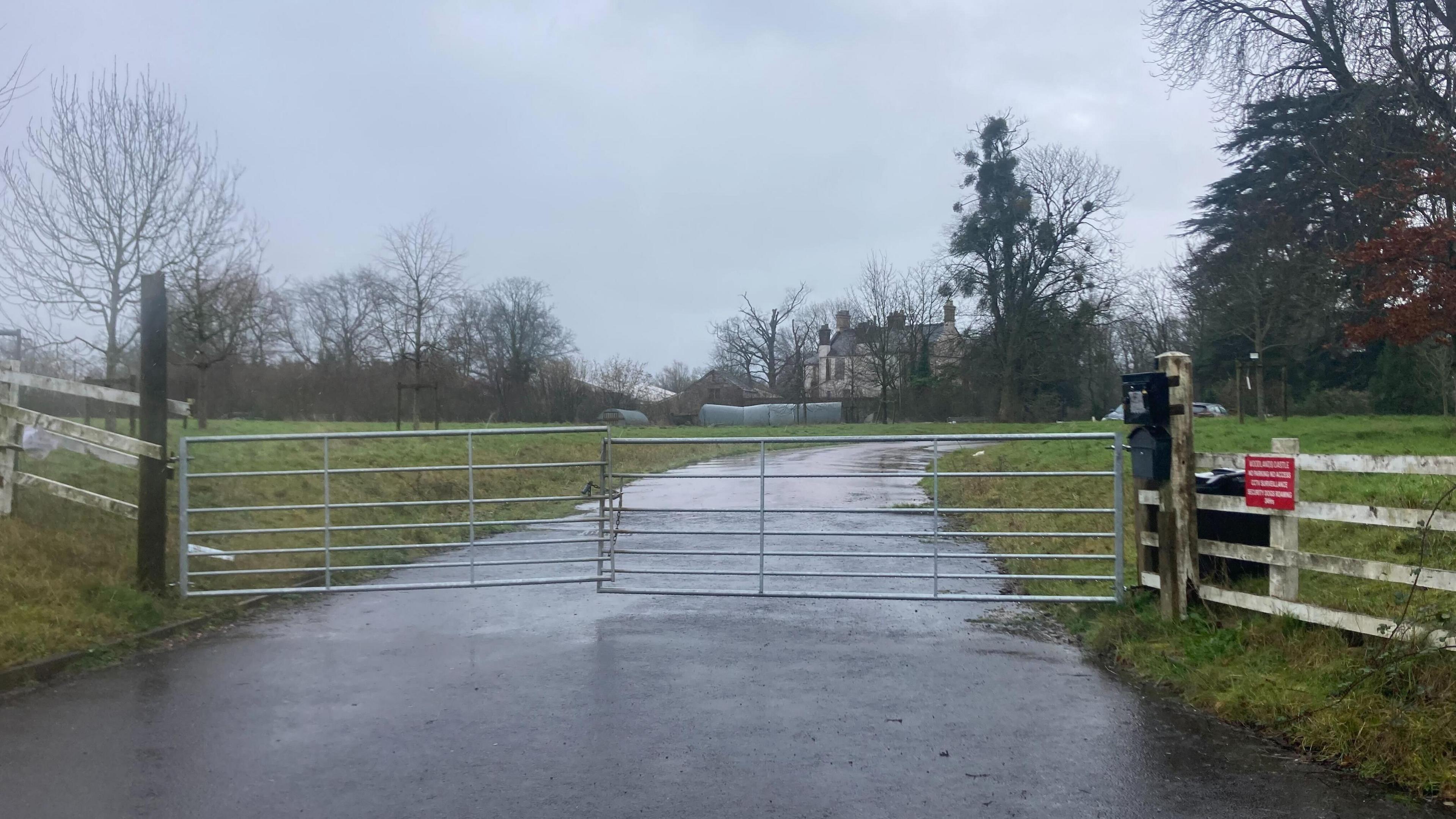 A set of metal gates closed over a driveway leading up to a big house surrounded by grass and trees. 