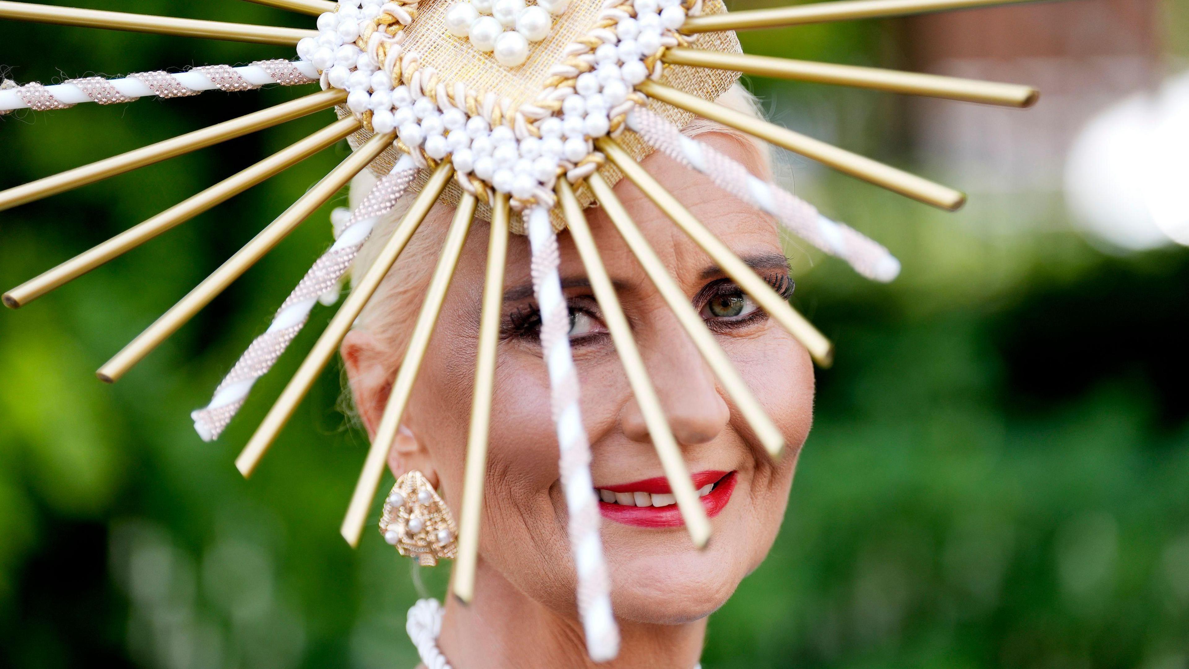 A woman smiles and looks at the camera. She has red lipstick on and large gold and pearl earrings. Her face is slightly obscured as she looks through the details of her fascinator hat, which is a gold headpiece covered in pearls, with a mixture of gold and pearl sticks coming out of it. The background is blurred and green.