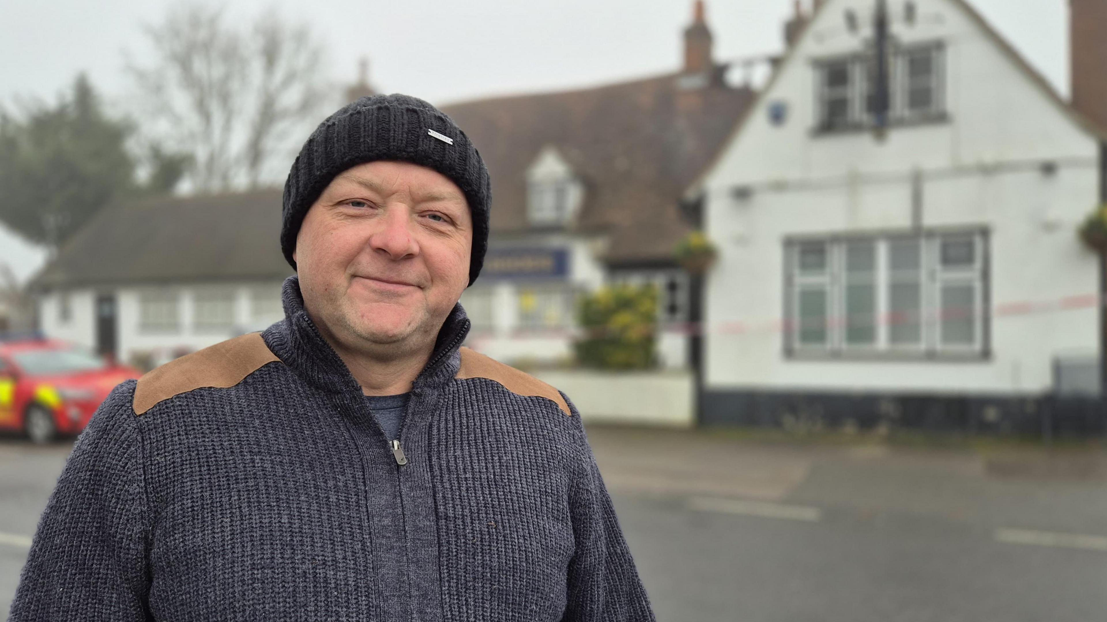 Paul Merritt is smiling at the camera and is wearing a black woolly hat and a dark blue cardigan. He is standing in front of the Three Horseshoes Pub