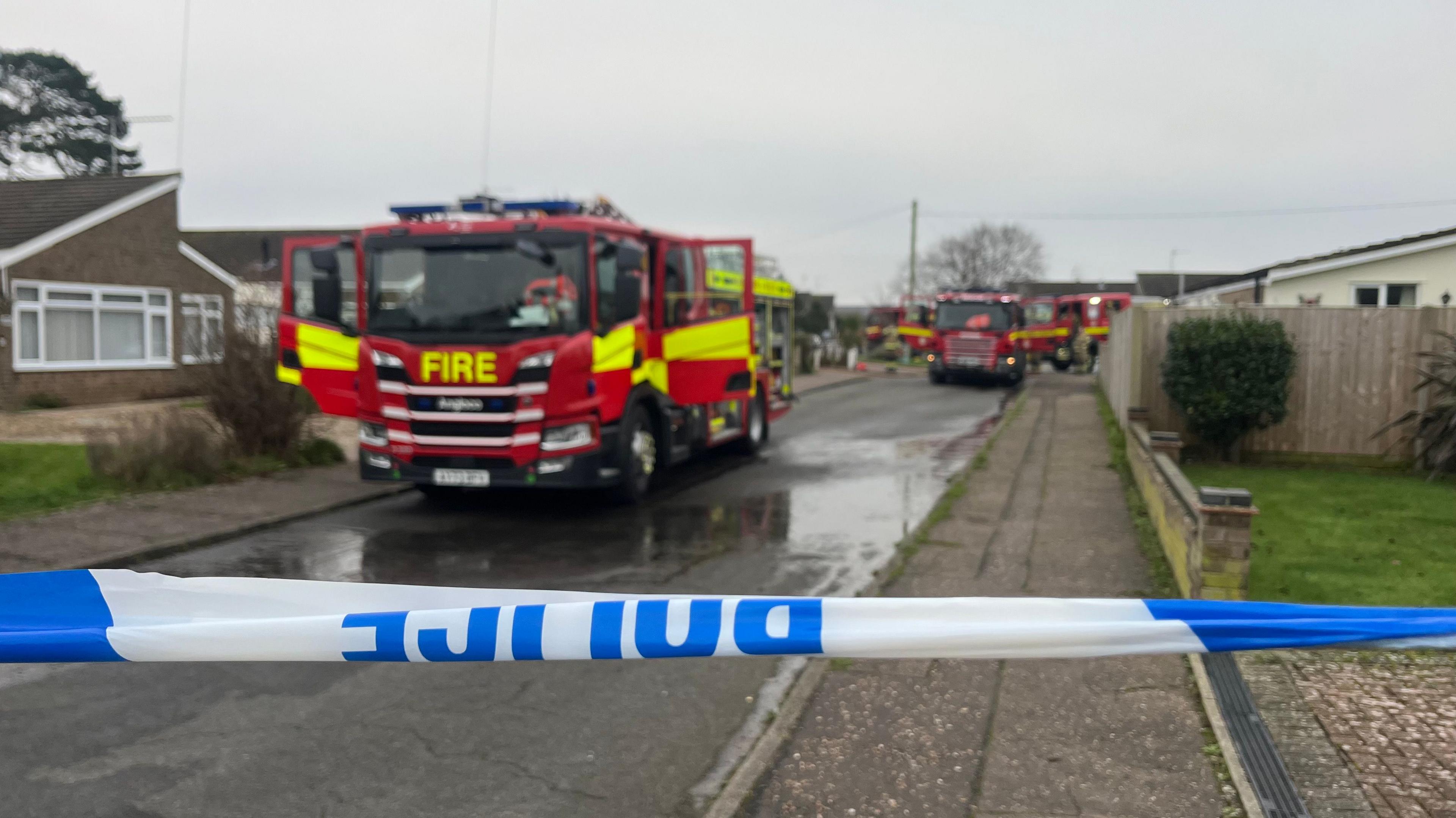 A number of fire appliances can be seen parked down a road. In front of the fire engines is a blue and white police tape to cordon off the area. 