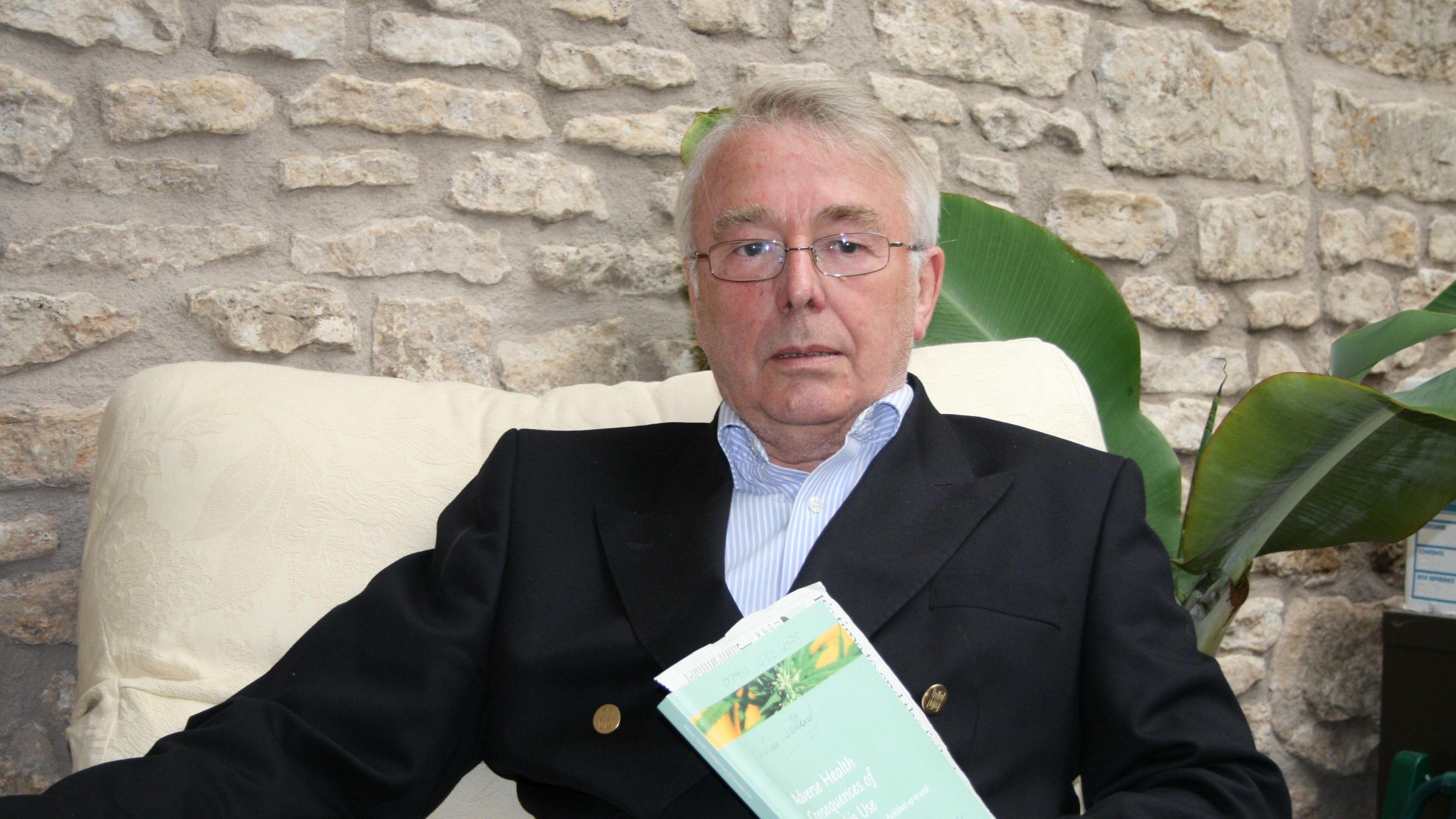 David Raynes is sat on a white chair, holding a brochure in his hand, wearing a navy suit jacket and a blue and white striped shirt.