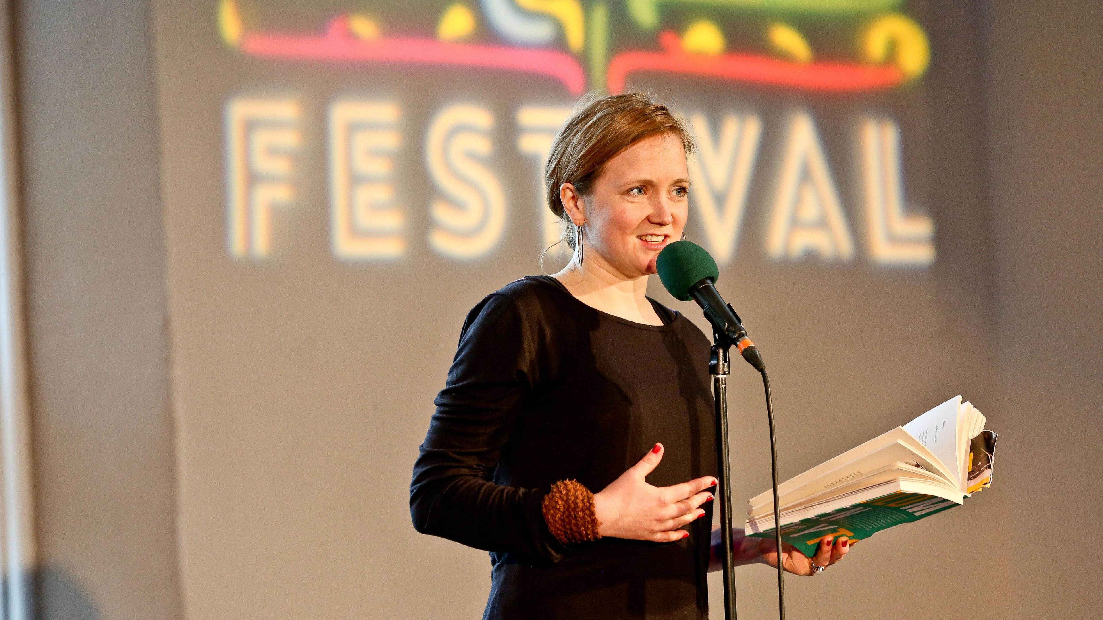 McNish stands at a mic with a book in her hand in front of a sign saying Festival