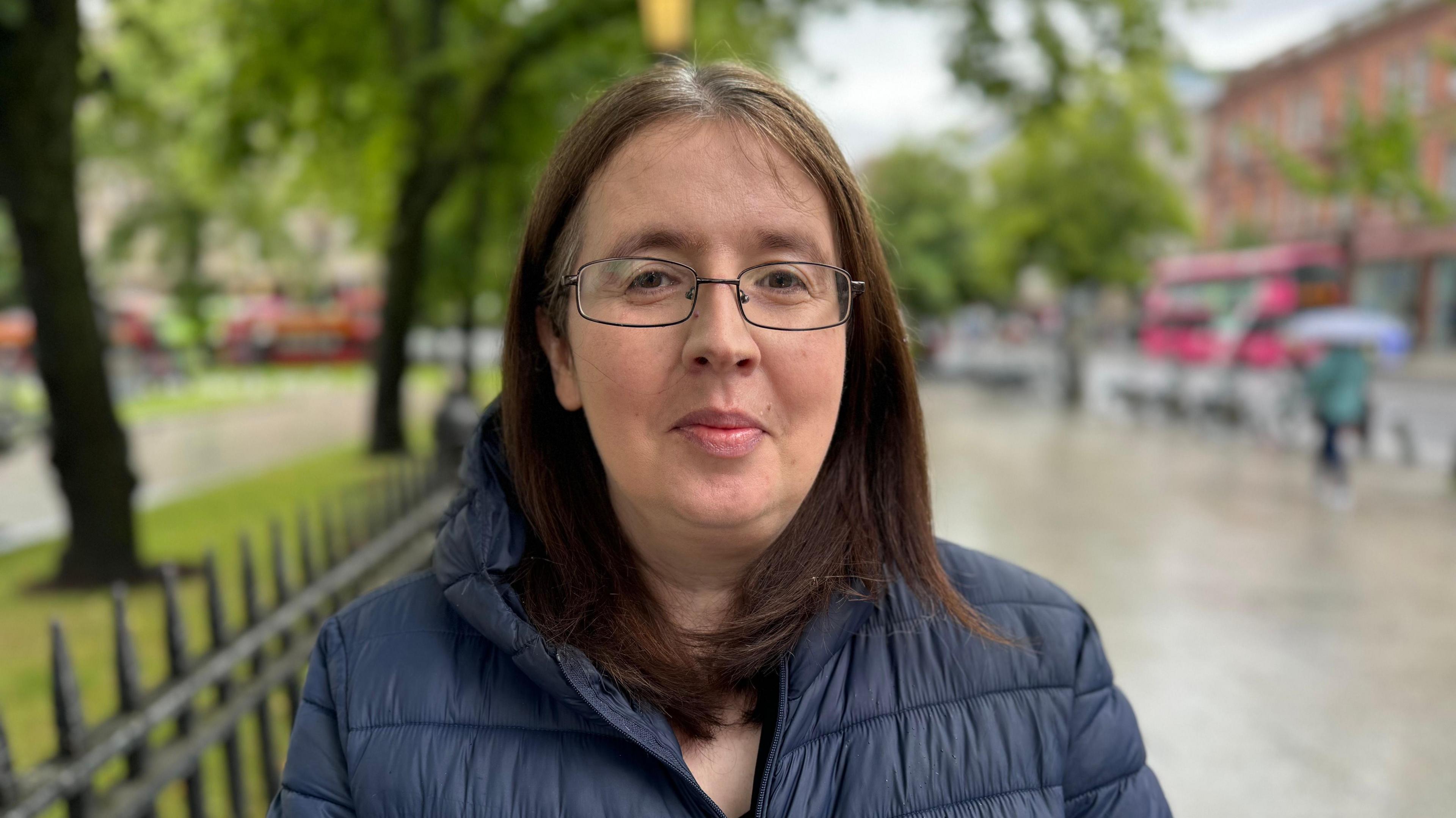 A woman with brown hair and glasses looking into the camera. She is standing on a city centre street wearing a navy blue quilted jacket.