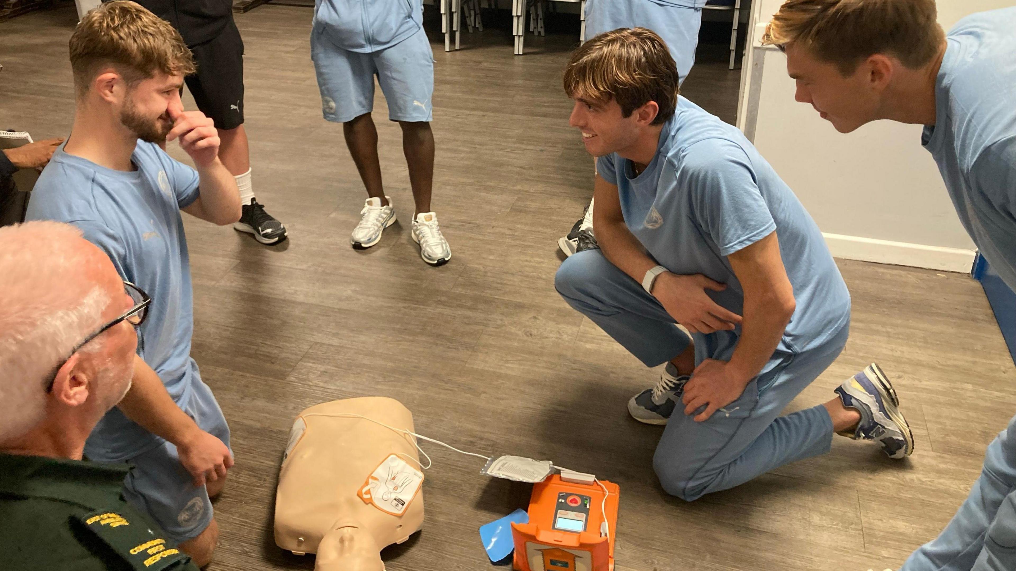 Men in blue kits surround a dummy used for CPR training as an emergency worker looks on.