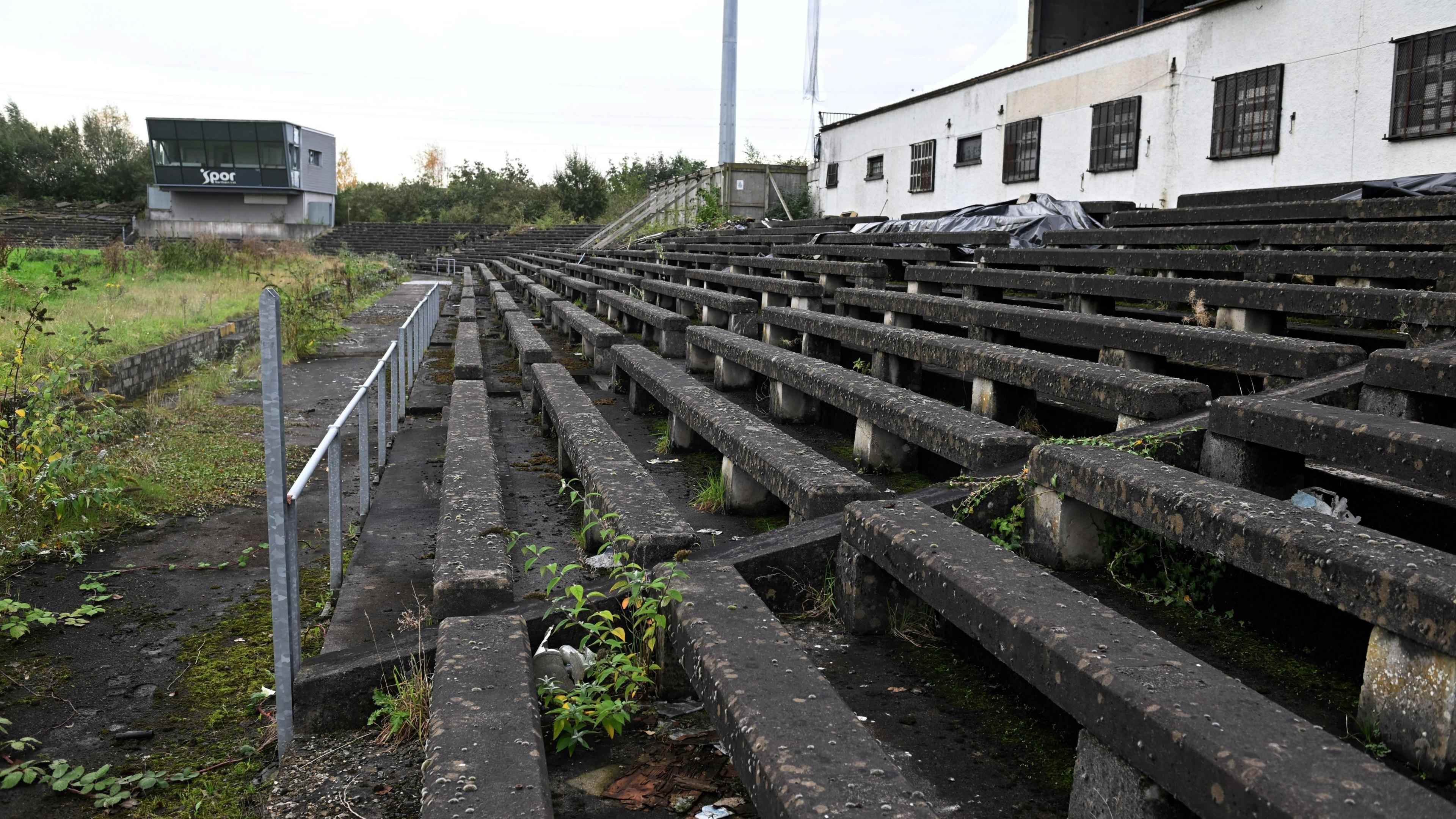 A close-up of the old concrete terraces of Casement, with weeds growing between them. A white building is behind them on the right of the picture and another empty grey two-storey building is on the left.